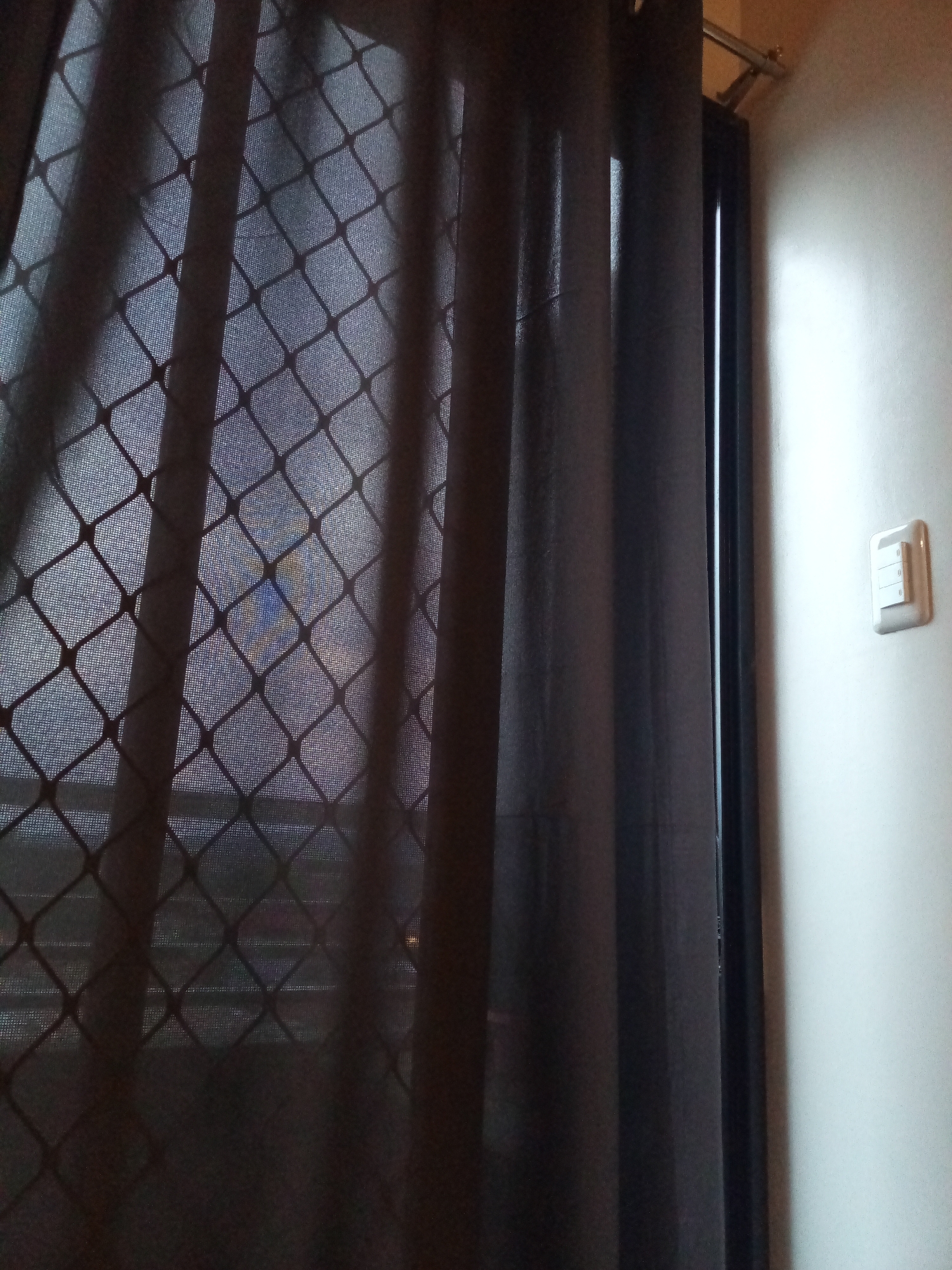 A close-up, indoor shot shows a dark curtain partially covering a window, the window's patterned screen visible through the curtain's folds. Adjacent to the window, a section of a light-colored wall is visible, featuring a light switch with three switches. The location is potentially in Davao City, Philippines.