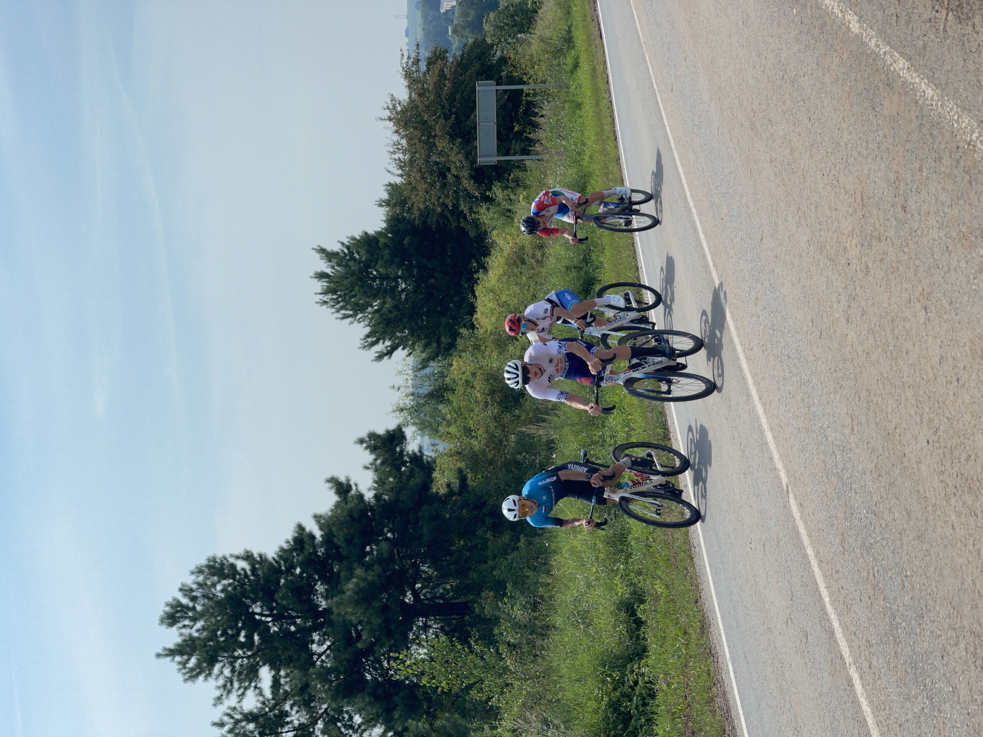 Four cyclists, wearing cycling attire and helmets, are riding on an asphalt road. The cyclists are in single file, with the lead cyclist in a blue and white jersey, the second in a white and red jersey, the third in a white and blue jersey, and the fourth in a red, white, and blue jersey. They are positioned on the right side of the road, with a white line marking the lane. The road is bordered by a grassy verge on the left, with lush green vegetation and tall trees in the background. The sky is clear blue with some wispy clouds. A road sign is visible on the right side of the road in the background. The scene appears to be a road race or training ride taking place in an outdoor, rural environment.
Based on the provided information, the location is Perevoznoye, Russia.