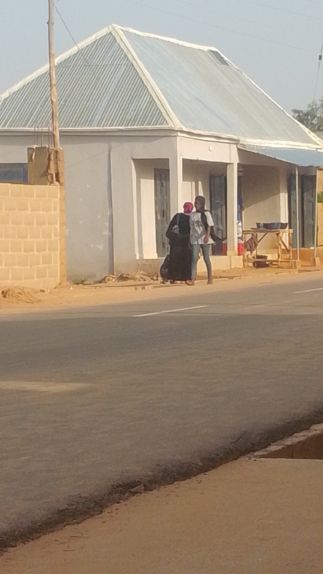 The image shows two people standing on a dusty roadside in front of a building. The building has a light-colored facade, a corrugated metal roof, and what appear to be shop entrances. To the left of the building is a brick wall. A wooden stall with items on display is visible to the right of the building.
The person on the left is wearing a dark, full-length garment and a red headscarf. They are carrying a large black bag. The person on the right is wearing a light-colored t-shirt with a graphic on it, blue jeans, and a headscarf. They appear to be interacting, possibly conversing. The ground is sandy and there is a paved road in the foreground. A utility pole is visible to the left of the building.
The lighting suggests it is daytime, likely late afternoon given the shadows. The overall scene appears to be in a somewhat arid or dusty environment. No text is visible in the image. The activity appears to be casual, with the two individuals paused on the roadside.