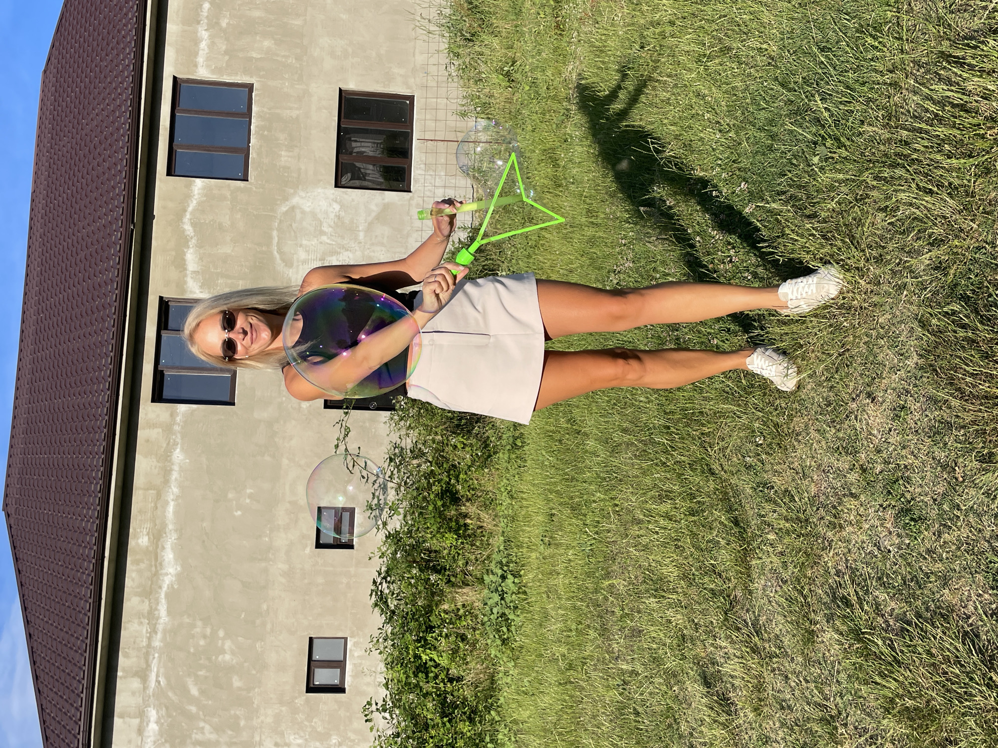 A young woman with blonde hair and sunglasses is playing with bubbles on a sunny day in Baranovka, Russia. She is standing on a grassy lawn in front of a light-colored, unfinished building with a brown roof. She is holding a green bubble wand and has a large iridescent bubble in her left hand. The woman is wearing a dark top and a light-colored skirt, and white sneakers. The sun is shining brightly, casting shadows on the grass. The overall mood is cheerful and playful.