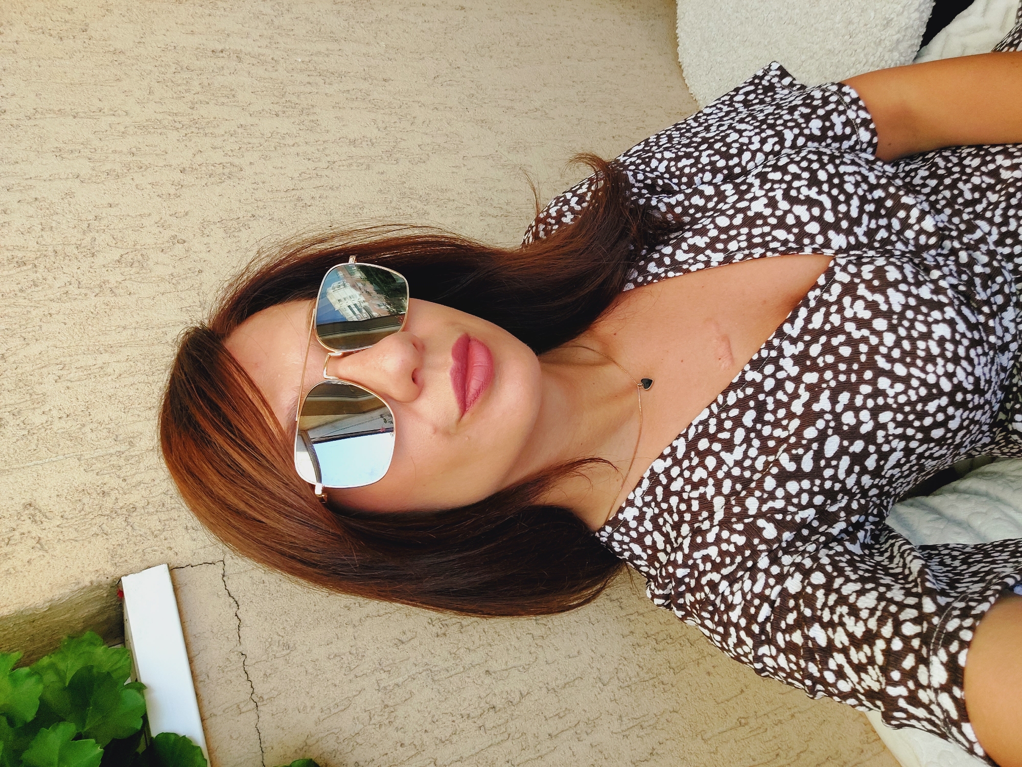 A woman is captured in a medium shot, looking towards the camera with sunglasses on. She has medium-length, reddish-brown hair and is wearing a brown and white spotted dress with a plunging neckline. A small, dark heart-shaped pendant hangs on a thin gold chain around her neck. A scar is visible on her upper chest. The background consists of a textured, light-colored wall with a crack running through it. To the left of the frame, there's a white ledge with green foliage. The lighting suggests an outdoor setting. The reflection in her sunglasses shows buildings and the sky. The image appears to be a selfie, taken from a slightly low angle. The woman is likely outdoors in Sofia, Bulgaria, based on the architectural style visible in the sunglasses reflection, which often features Soviet-era apartment blocks.