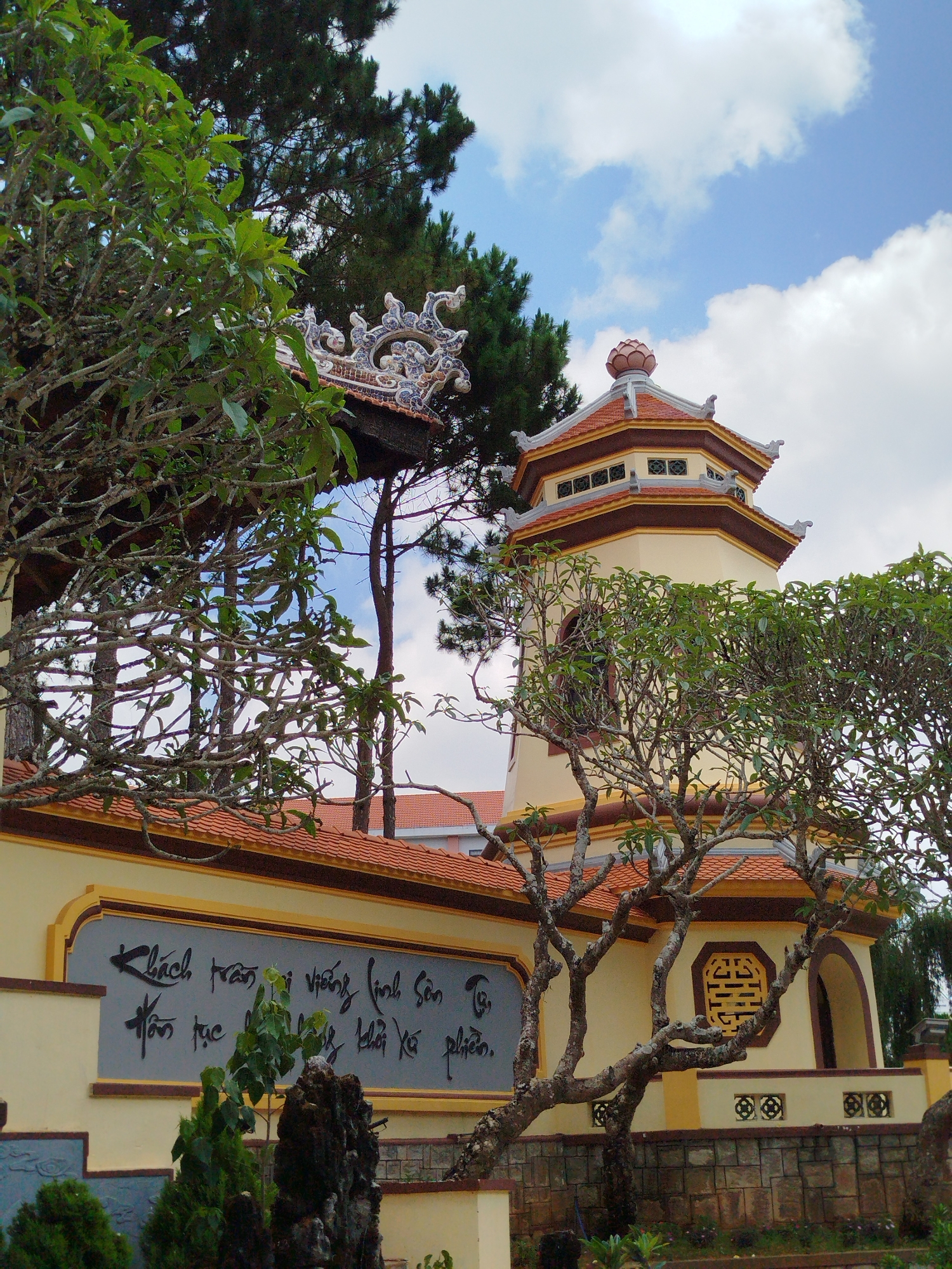 This image captures an architectural scene, likely a Buddhist temple or pagoda in Da Lat, Vietnam, set under a bright, partly cloudy sky. The central subject is a multi-tiered, octagonal tower with cream-colored walls, accented by dark brown trim and reddish-brown tiled roofs. A lotus-shaped finial crowns the tower's uppermost section. Ornate, light-colored ceramic decorations adorn the roof ridges of a building partially visible on the left.
The foreground features several trees with gnarled, intertwining branches, some bearing green leaves, partially obscuring the structures and adding a natural, aged feel to the scene. Taller, darker pine trees are visible in the background, suggesting a forested or mountainous environment typical of Da Lat.
A prominent feature is a grey rectangular panel on a cream wall, displaying elegant black Vietnamese calligraphy. The visible text appears to read:
"Khách trần ai viếng linh sơn Tục tấc"
"Hồn tục khôi xa phiền."
This translates to a sentiment often found in temples, inviting visitors to the sacred place to shed worldly worries. Below the calligraphy, a patterned stone wall forms the base of the structure. Further architectural details include an octagonal window with a decorative lattice pattern resembling a Chinese character and smaller circular grilles below.
The scene is illuminated by daylight, indicating good weather. No people are visible, lending a serene and contemplative atmosphere to the traditional setting.