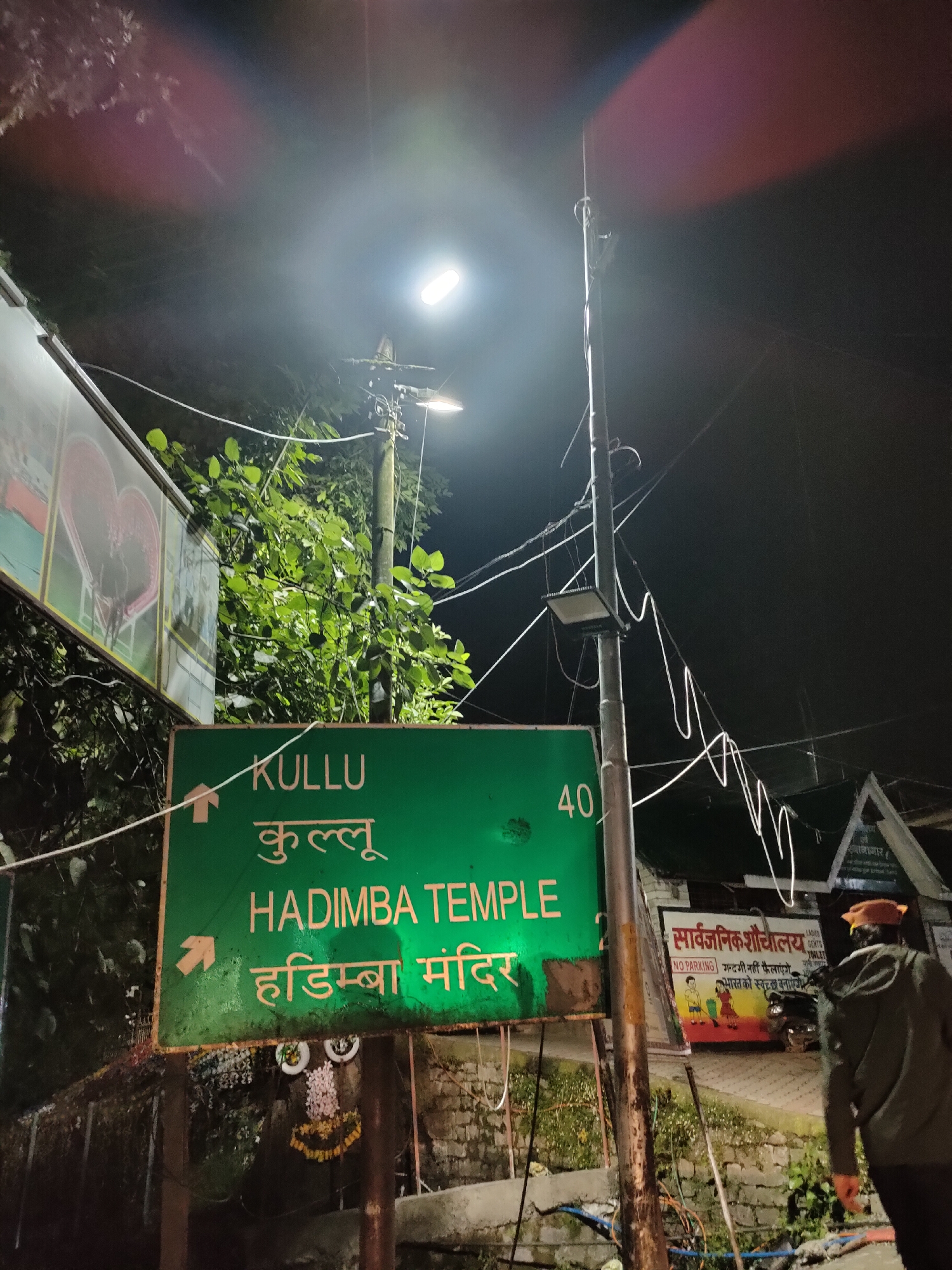 This image depicts a night scene in Manali, India, focusing on a directional sign for Kullu and Hadimba Temple. The sign is green with white and yellow text, indicating directions to "KULLU" and "HADIMBA TEMPLE." The text is in both English and Hindi script. An arrow points left for Kullu, and another arrow points left for Hadimba Temple. To the right of the sign, the number "40" is visible, likely indicating a distance.
In the background, a tall utility pole with a bright street light illuminates the area. Several wires are strung across the pole and the scene. Behind the sign, a smaller, triangular sign displays text in Hindi, possibly related to a public facility. A person wearing a dark jacket and a cap is walking away from the camera towards the right side of the image, partially obscuring a colorful advertisement board. The environment is somewhat overgrown with foliage, and the ground appears to be a mix of paved surfaces and rough terrain. The overall atmosphere is typical of a street in a tourist town at night, with artificial lighting creating strong contrasts.