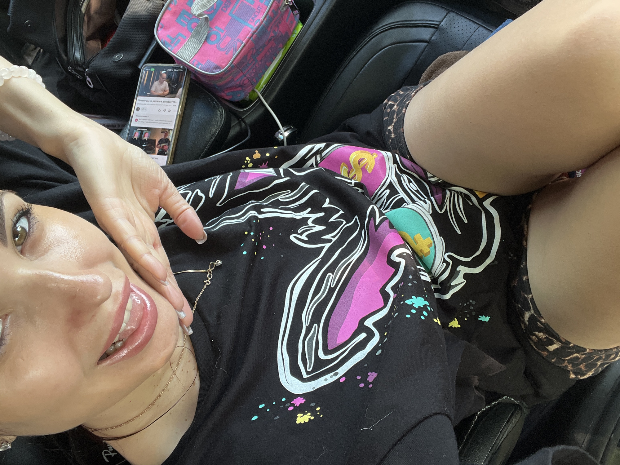 A young woman with dark hair and brown eyes, wearing a black t-shirt with a colorful graphic and leopard print shorts, is taking a selfie in the car. She is smiling with her tongue sticking out, and her hand is resting on her face. A phone, a pink bag, and other items are visible in the background. The car appears to be parked, possibly in a city, though the specific location is unknown.