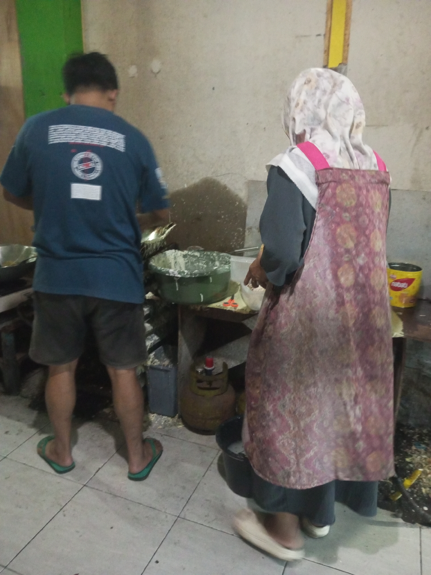 In a dimly lit, cluttered room that appears to be a kitchen or food preparation area in Andongsili, Indonesia, two individuals are engaged in an activity, possibly cooking or preparing food. A man, viewed from the back, wears a dark blue t-shirt with a logo and a white rectangular print on the back, and dark shorts. He stands near a large, green basin filled with what looks like food ingredients, and a metal bowl. To his right, a woman, also seen from behind, is wearing a gray long-sleeved shirt and a patterned pink and maroon apron over a light-colored hijab. She is standing near a counter with a yellow container. The room has a concrete wall and tiled floor. A gas cylinder is visible behind the woman. The overall impression is one of a busy, functional workspace.