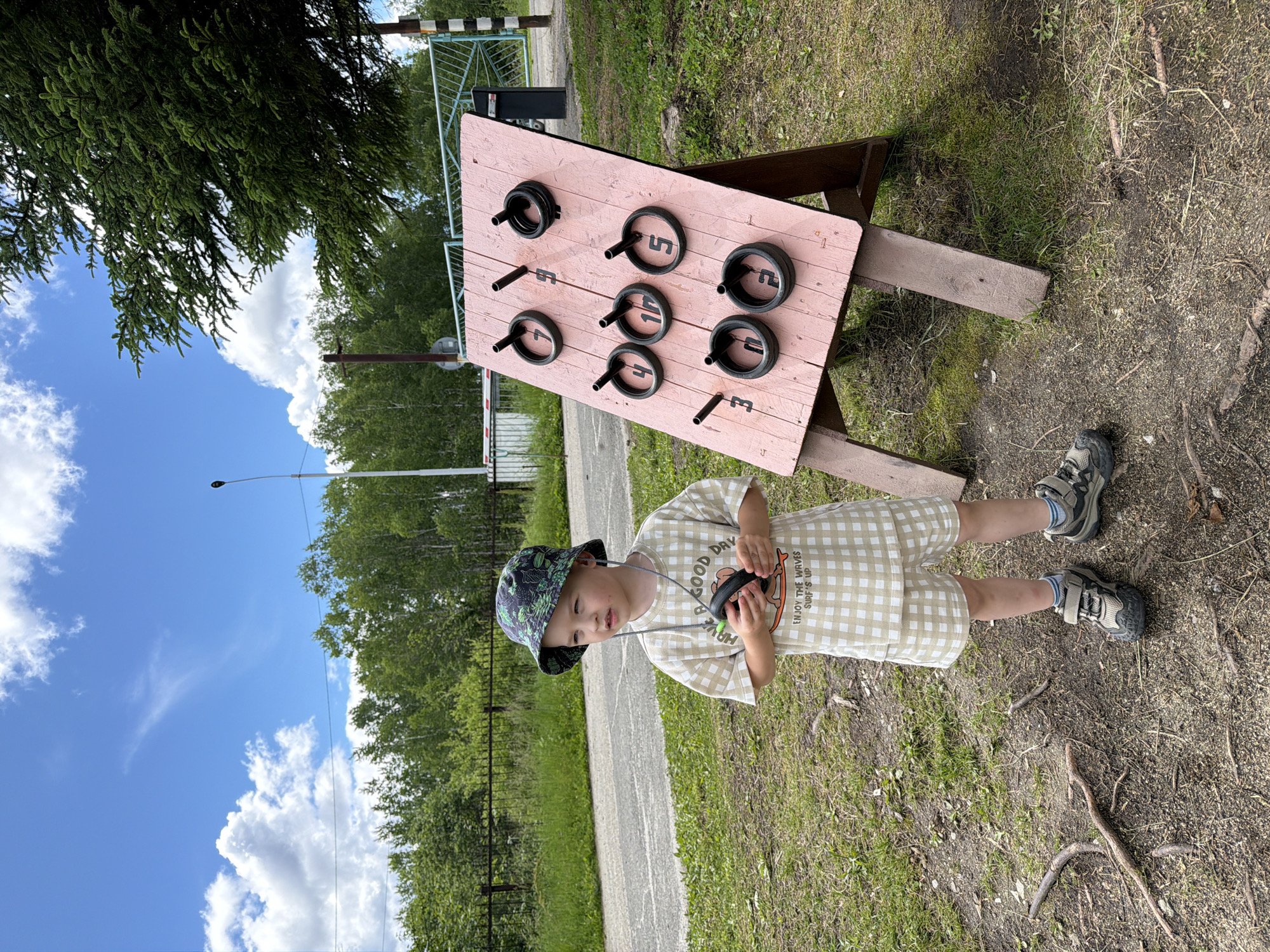 A young boy stands in a grassy area next to a ring toss game in Kul'dur, Russia. The boy, wearing a bucket hat and a checkered t-shirt and shorts, holds a black ring in his hands. He is looking to his right. The ring toss game is set up on a wooden easel and has a pink board with pegs and rings attached. The background features trees, a road, and a blue sky with white clouds. A metal fence and a gate are visible in the distance.