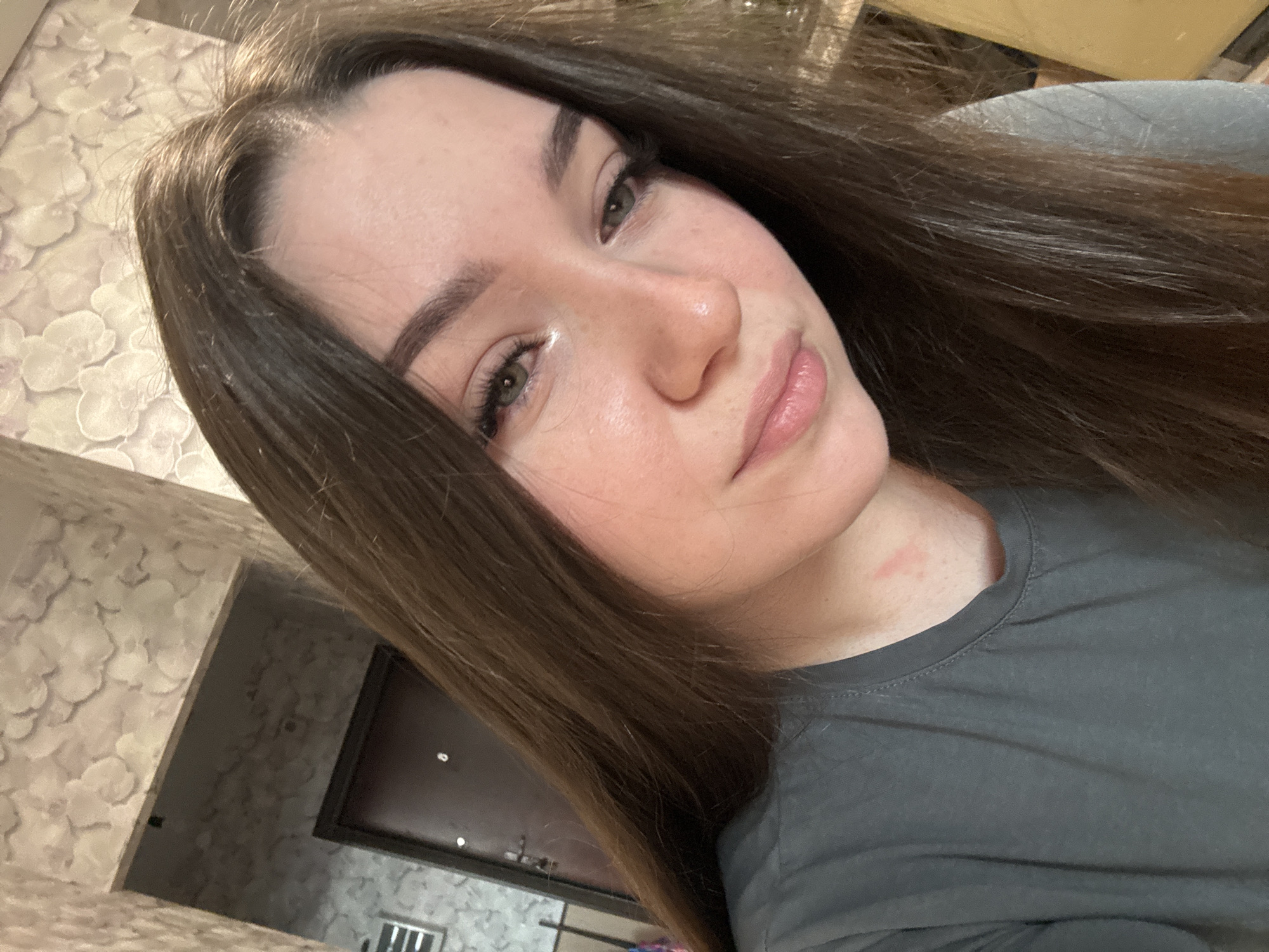 A selfie captures a young woman with long, brown hair and light-colored eyes. She is wearing a dark gray t-shirt. The background appears to be an indoor setting with patterned wallpaper and a doorway. There is a reddish mark on her neck, just below her ear. The image was taken in Zarechnaya (Gamovskoe S/P), Russia.