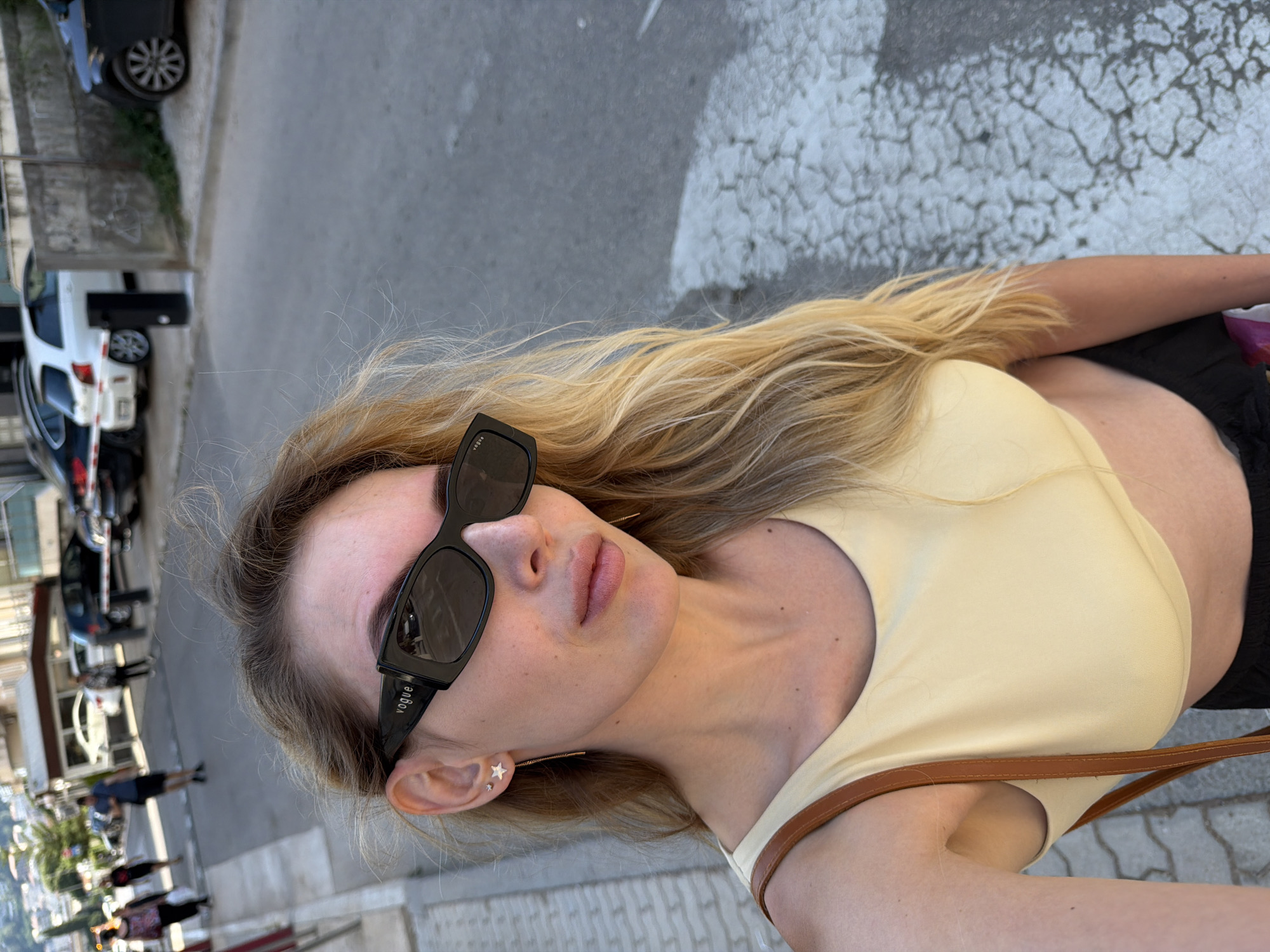 The image captures a young woman with blonde hair and sunglasses, wearing a light yellow top and black bottoms. She is leaning back, her head tilted to the side, and appears to be taking a selfie. Her facial expression is relaxed, and the lighting suggests it is daytime. She has star-shaped earrings and a subtle, delicate necklace or chain visible around her neck. The background shows a street scene with cars parked along the side of the road and a crosswalk. The architecture visible in the distance suggests an urban or town setting, consistent with the provided location context of Budva, Montenegro. The sunglasses have the word "VOGUE" printed on them.