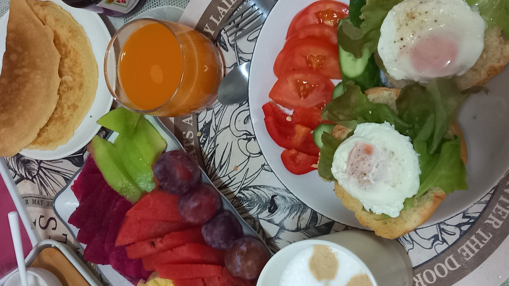 A close-up, eye-level shot captures a delectable breakfast spread on a table, likely at a restaurant or hotel in the Ko Pha-ngan District of Thailand. The foreground is dominated by a white plate featuring two open-faced sandwiches. Each sandwich consists of a toasted bread base topped with fresh green lettuce and a perfectly poached egg, seasoned with pepper. Next to the sandwiches, sliced tomatoes and cucumber add a vibrant splash of color.
To the left, a white mug holds a frothy, light-brown beverage, possibly coffee or a latte, with a decorative pattern on its surface. Behind the mug, a small white bowl with a white spoon is visible, hinting at other breakfast accompaniments.
The mid-ground showcases a colorful fruit platter arranged in a white dish, with slices of watermelon, dragon fruit, and melon. A few dark red plums are also scattered on the plate. Adjacent to the fruit, a clear glass is filled with a bright orange beverage, likely fruit juice.
In the background, a stack of golden-brown crepes or pancakes rests on a white plate. A small jar of what appears to be jam, with a purple label, is positioned to the right of the pancakes. The table is covered with a patterned placemat, featuring a black and white sketch of leaves and flowers, with some text in English. The overall impression is one of a healthy and appealing breakfast, set against a tropical backdrop.