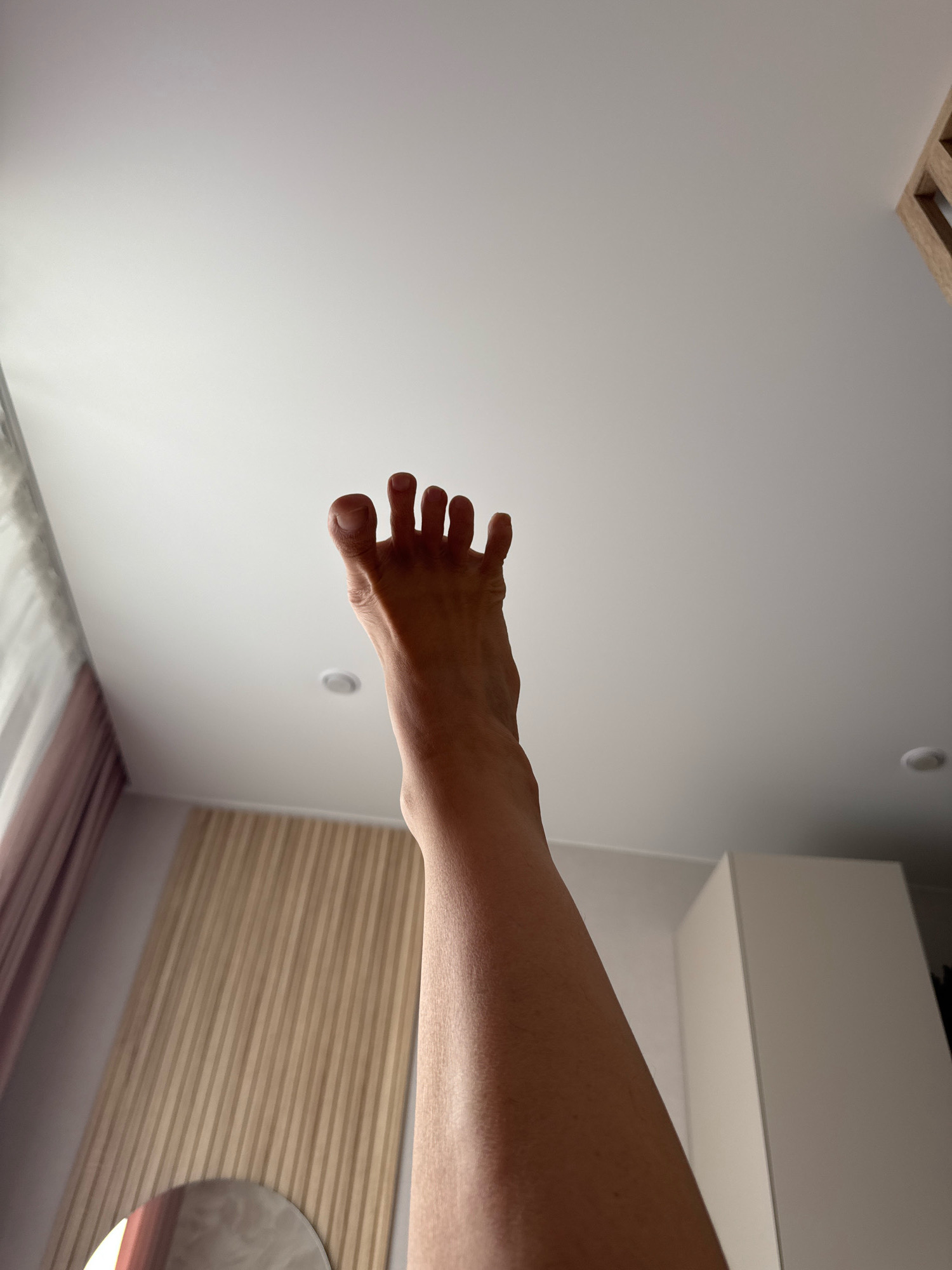 A person's left foot is extended upwards against a white ceiling. The toes are spread apart. The leg is visible from the ankle to mid-calf. In the background, on the left, is a window with sheer curtains. To the right of the foot, there is a slatted wooden wall panel. Further to the right, there is a tall, white cabinet. The scene appears to be indoors, possibly in a bedroom or living area. There is no information in the image to suggest this location is in Simferopol, Ukraine.
