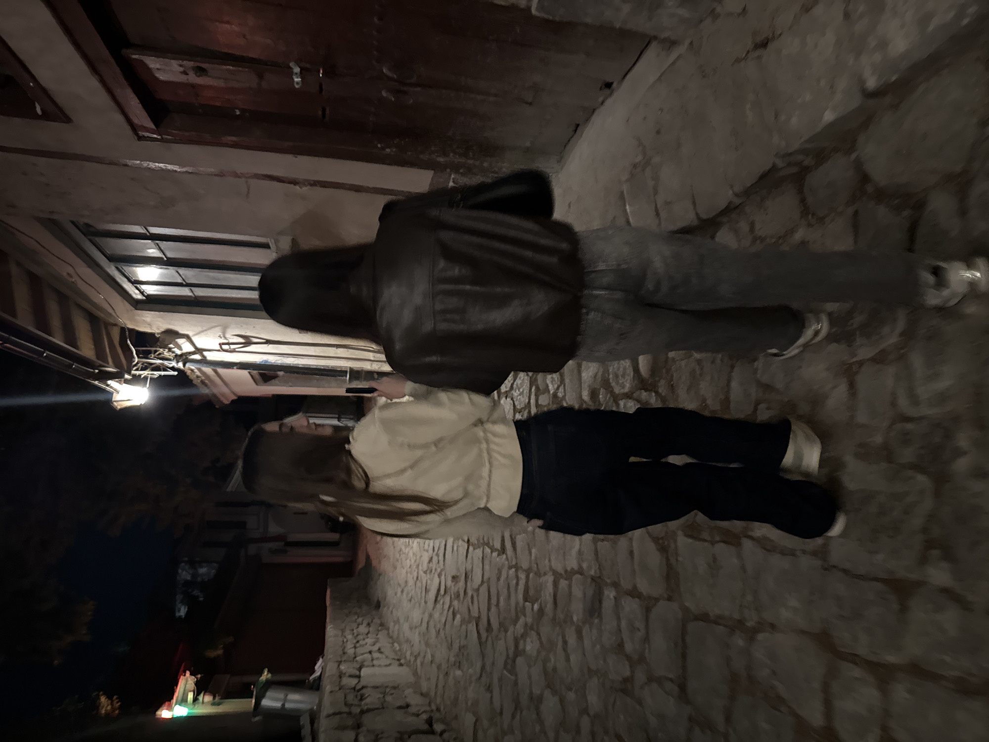 Two people are walking on a cobblestone street. The person closest to the camera is wearing a light-colored jacket and dark pants. The person further away is wearing a dark leather jacket and light-colored pants. The street is narrow and lined with buildings on both sides. The time is night. The location appears to be in the Old Town of Plovdiv, Bulgaria.