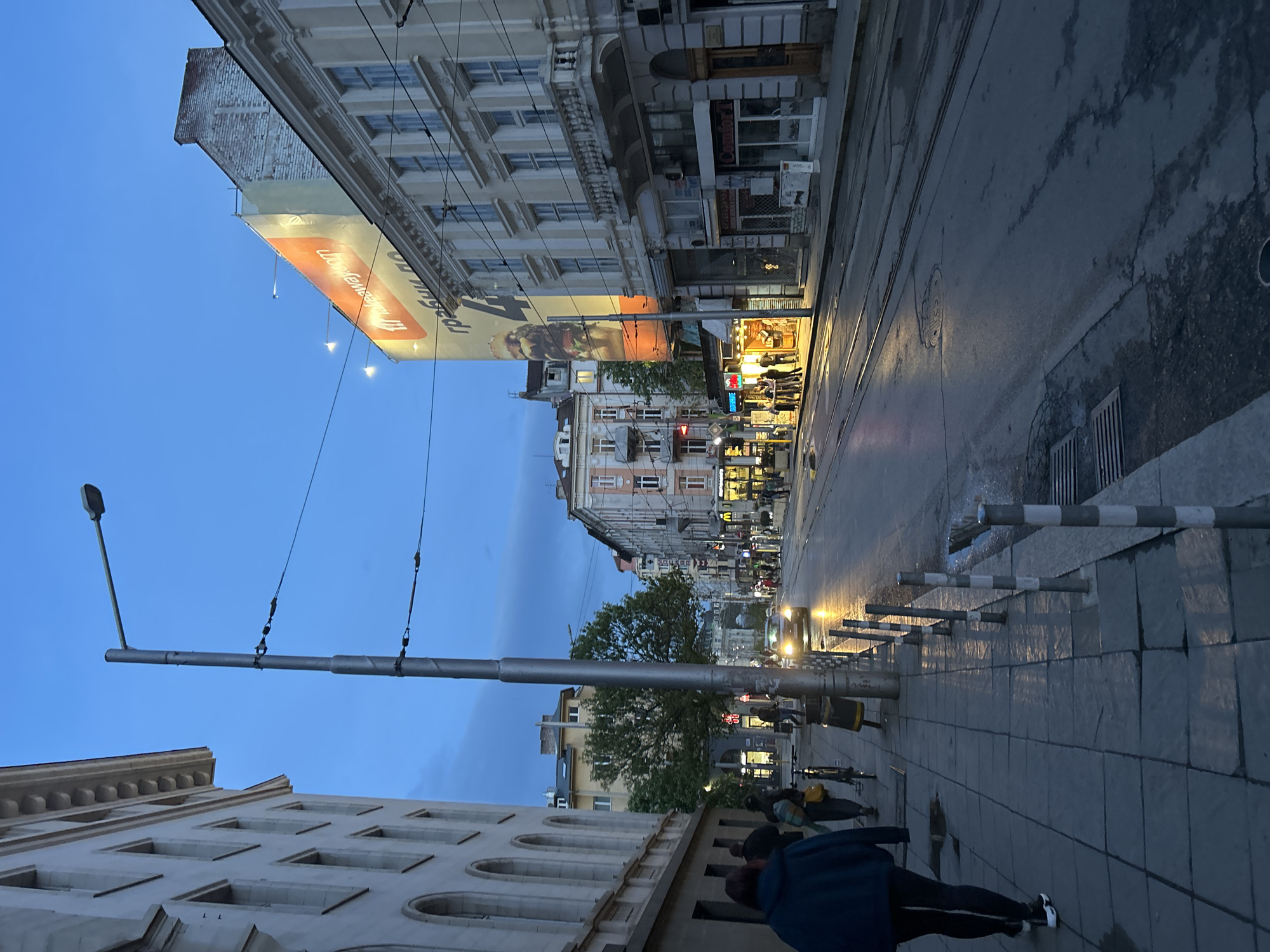 The image shows a street scene in Sofia, Bulgaria, captured at dusk. The street is lined with buildings, displaying a mix of architectural styles. A large advertisement is prominently displayed on one of the buildings. A row of traffic control poles borders the sidewalk. People are walking along the sidewalk, and a car is visible on the wet street. The street lamps are lit, reflecting on the wet road surface. Overhead electrical lines are visible. The sky is a deep blue.