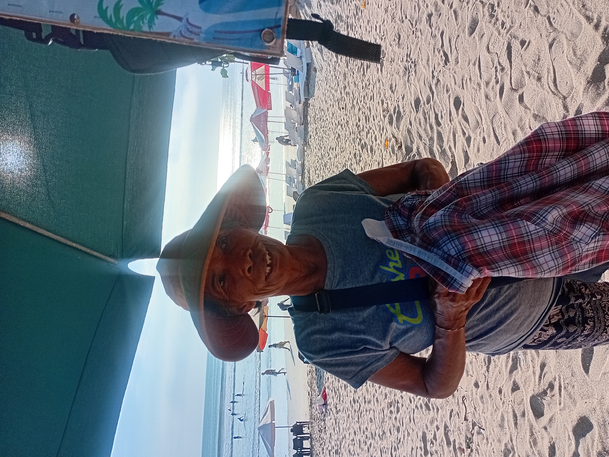 A portrait captures a woman wearing a wide-brimmed hat and a gray t-shirt standing on a sandy beach in Kuta, Indonesia. The woman is holding a red and white plaid shirt. In the background, beach umbrellas and lounge chairs are visible, along with people walking and swimming in the ocean. The sun is shining brightly, creating a glare on the water. The scene suggests a tourist-oriented beach environment.