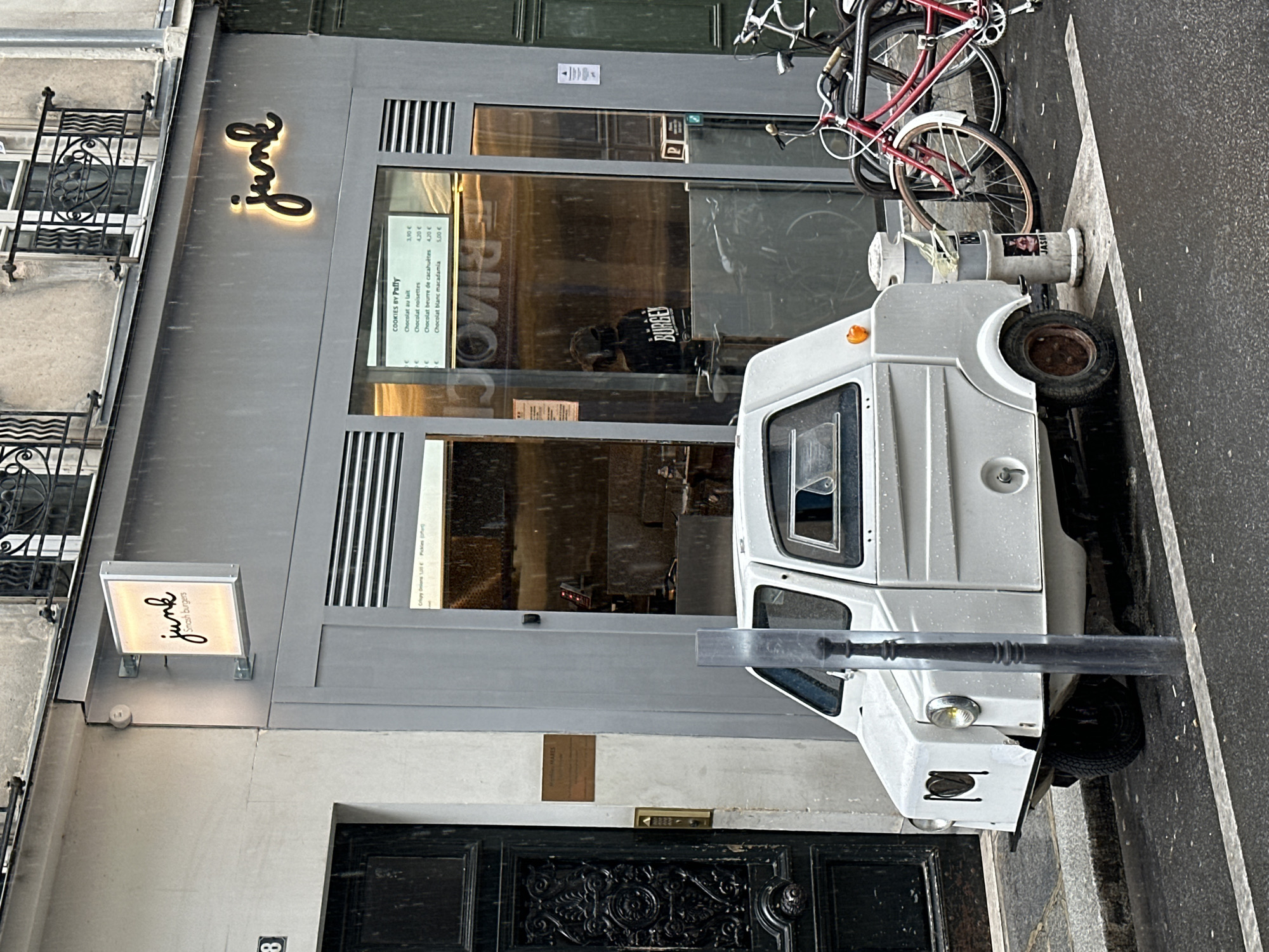 This is an exterior shot of a business front in Paris, France. A white, small, electric utility vehicle is parked on the street. A bicycle is parked behind the vehicle. The building façade has a grey finish. "Junk" is illuminated in stylized font above the storefront. A smaller sign adjacent to the entry door also reads "Junk Smash burgers." The number "8" is visible on the building near the bottom left corner. The menu board for the establishment is visible inside the glass, advertising "Cookies by Puffy." The front door of the building is black with ornate detailing. A bollard is visible in front of the utility vehicle.