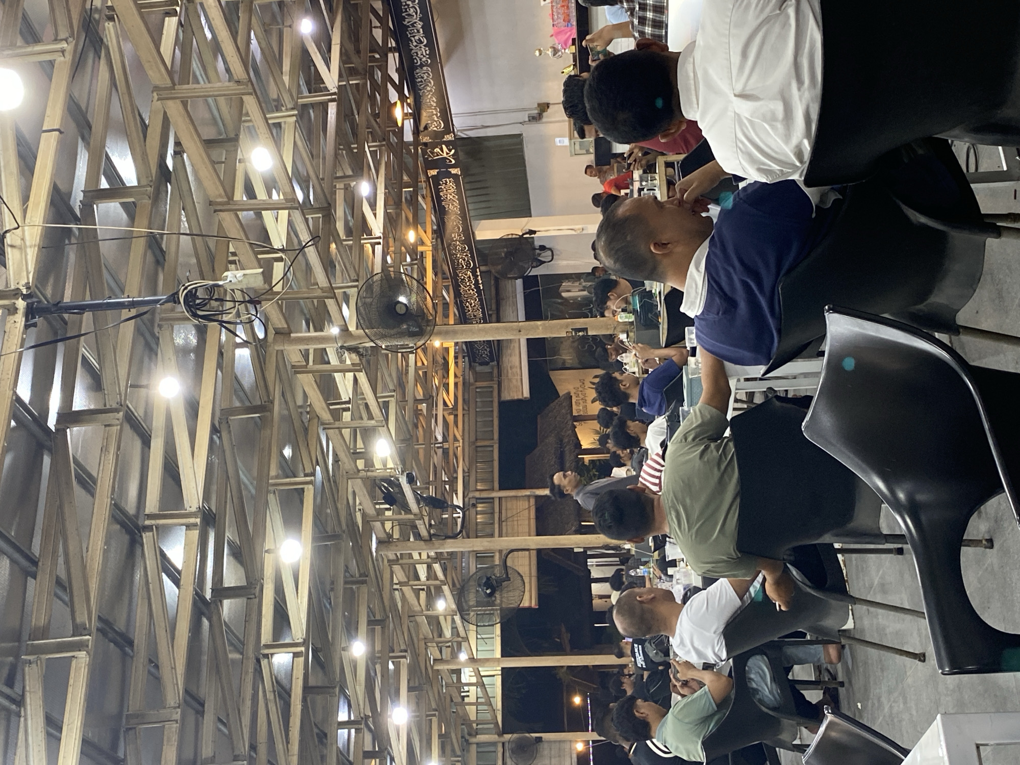 The image depicts a crowded indoor setting in Lhokseumawe, Indonesia. Many people are seated on black chairs, seemingly engaged in conversation. The building's structure features a wooden framework with numerous light bulbs illuminating the area. Several electric fans are mounted to the wooden beams. A banner with Arabic script is visible above the people. The general atmosphere suggests a social gathering.