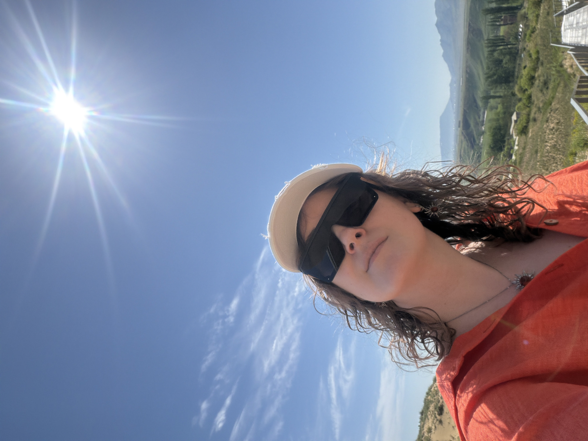 A medium shot taken from a low angle shows a woman wearing a pair of dark sunglasses, a light-colored hat, and an orange shirt. The woman is angled in a way that the viewer sees mainly her face and upper chest. She has curly hair. The background is a clear blue sky with a bright sun in the upper left and some sparse white clouds. Behind the woman, there is a green and brown landscape with some hills in the distance. The woman is on an observation deck. The location is Unknown, Unknown.