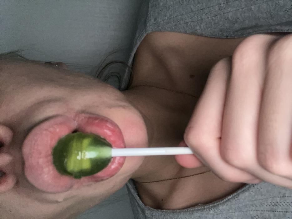 A close-up shot shows a person holding a green lollipop in their mouth. The lollipop's white stick is visible. The person's lips are prominent, and their hand grips the lollipop stick. Part of their face, neck, and gray shirt are also visible. A thin necklace adorns their neck. The image is slightly tilted, giving an unusual perspective.