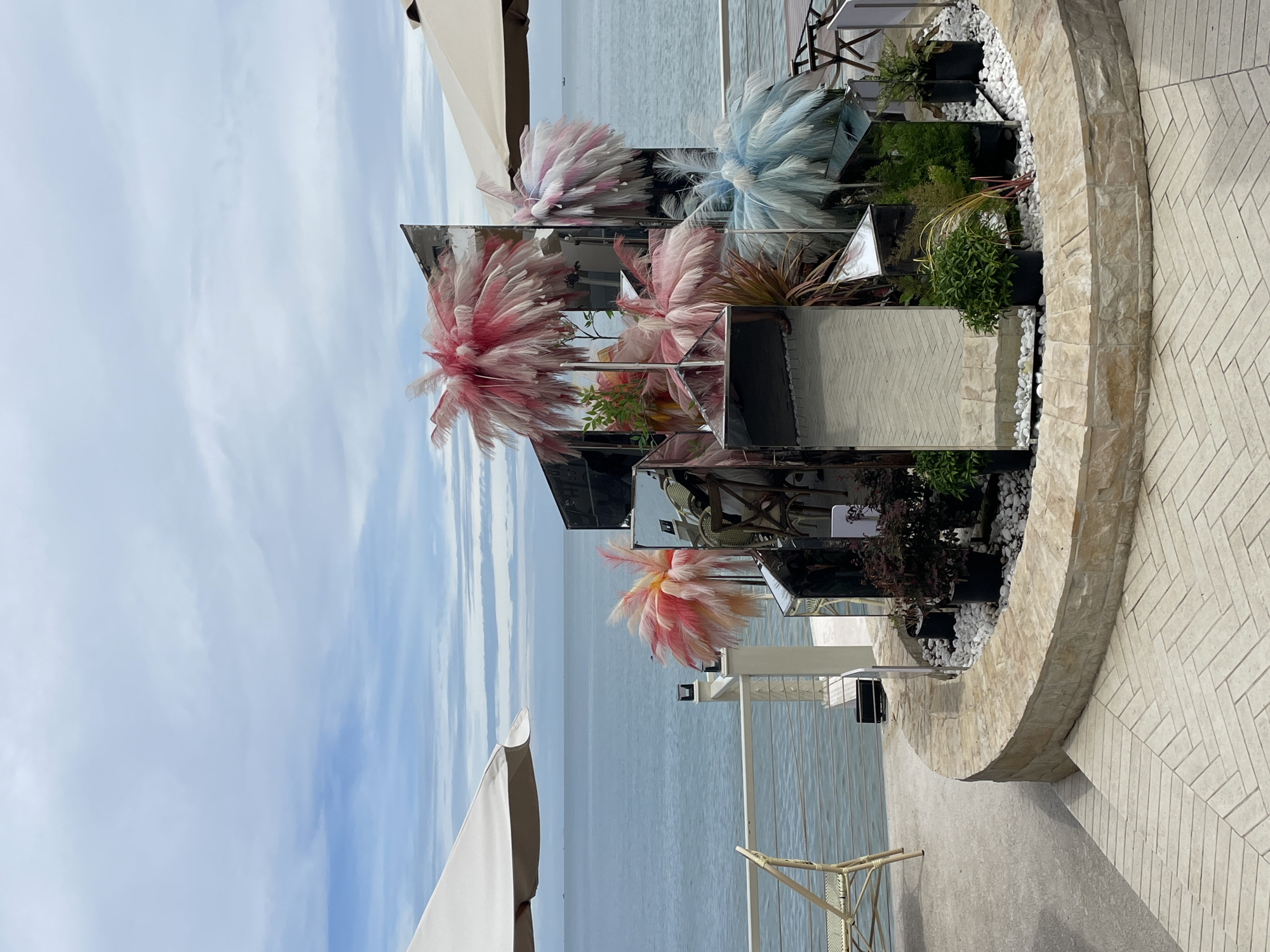 The image shows a decorative outdoor setting in Phu Thuy Ward, Vietnam. A tiered display of reflective square panels stands within a semi-circular planter outlined with light-colored stone. Within the planter are small plants in dark pots, surrounded by white gravel. Decorative feather plumes in shades of pink, white, and blue are interspersed among the reflective panels. The backdrop includes a view of the ocean and a sky with light, wispy clouds. Portions of a white umbrella and outdoor furniture can also be seen. The ground is paved with herringbone-patterned tiles.