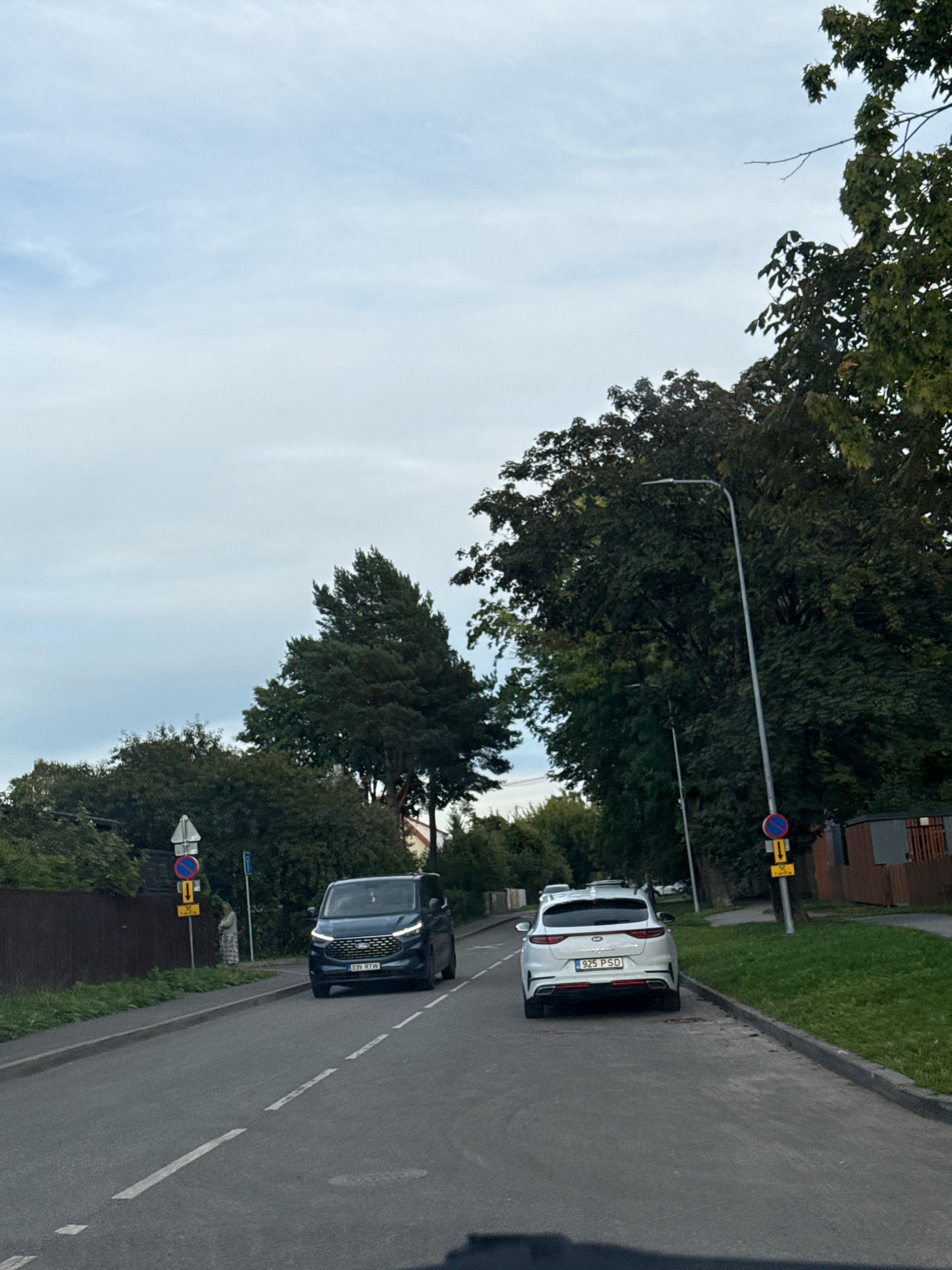 The image shows a street scene in Tallinn, Estonia. The road has dashed white lines indicating lanes. Two cars are visible: a dark blue minivan on the left and a white hatchback on the right. The minivan has the license plate "B39 RTW". The white hatchback has the license plate "925 PSD". There are trees and buildings lining the street. Traffic signs are present, including a "no stopping" sign and a sign with a yellow arrow. A person is standing on the sidewalk to the left of the minivan, wearing a patterned dress and a light-colored scarf. The sky is overcast.