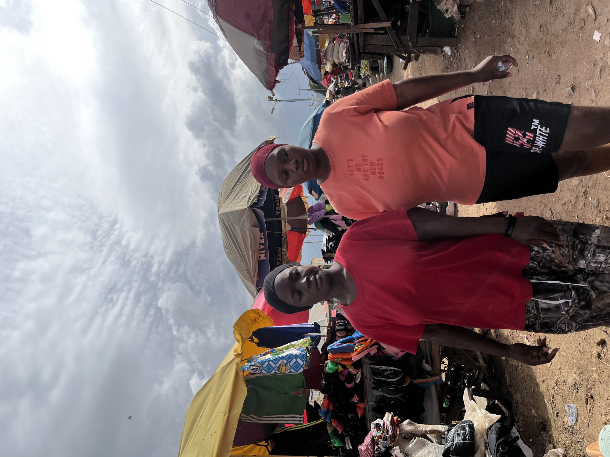 The image shows two young women walking in an outdoor market, likely in the city of Rafin Par, Nigeria. The market appears to be a bustling area with various stalls and vendors. In the foreground, the woman on the left wears a red t-shirt and patterned pants. The woman on the right is wearing a light orange t-shirt with text that reads "LET'S GO AND TRY NEW ROADS" and black shorts with a white graphic logo. Both women are wearing headscarves. Behind them, market stalls are visible, adorned with colorful fabrics, clothing, and what appear to be shoes or sandals. Several umbrellas are set up to provide shade. The ground is a dusty, reddish-brown earth, typical of many Nigerian environments. The sky above is overcast with dramatic clouds. The overall scene depicts a typical day in a Nigerian open-air market, with people engaged in commerce and daily activities.