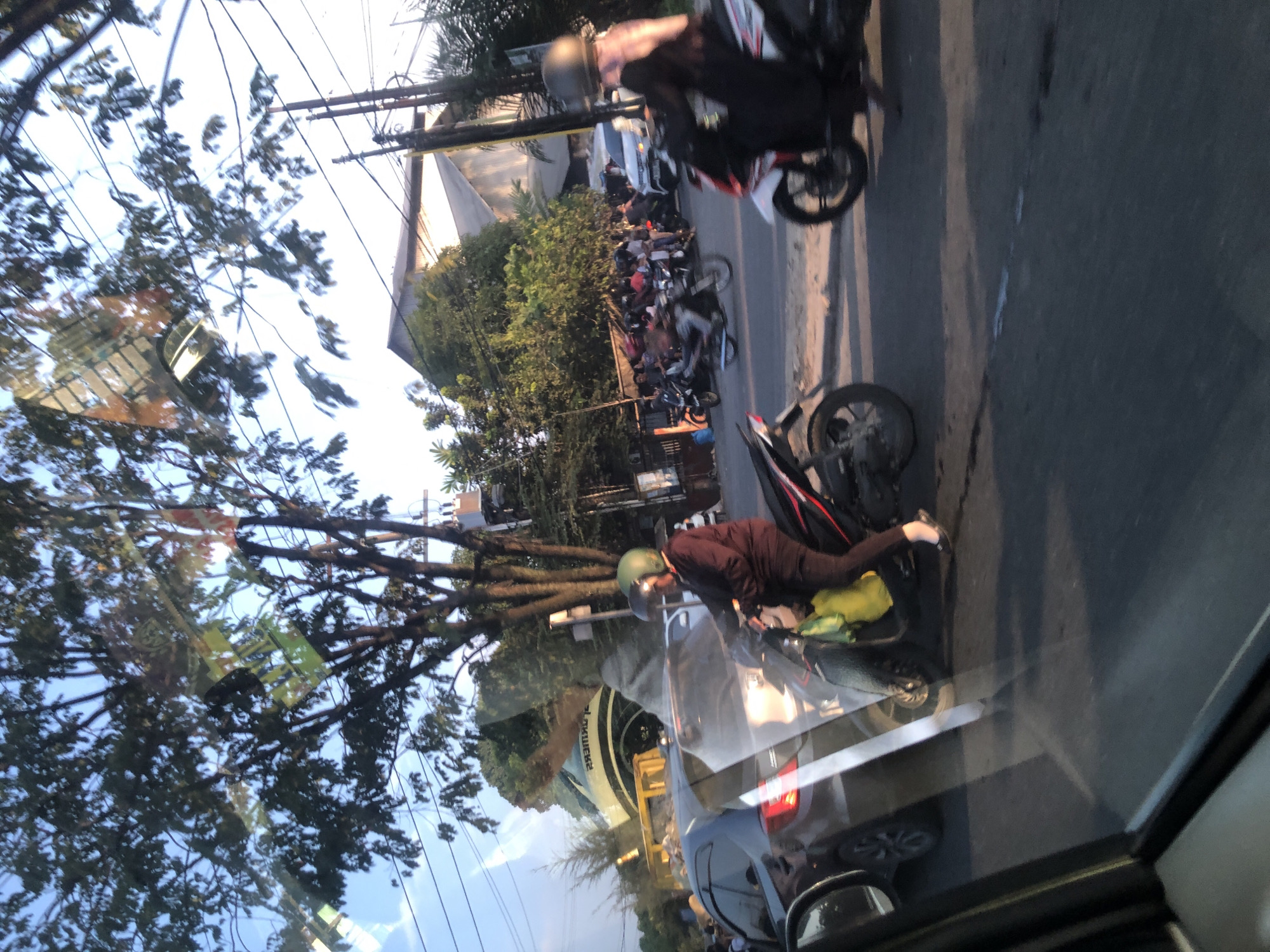 The image captures a street scene in the City of Medan, Indonesia, likely during daylight hours with a low sun angle casting long shadows. The perspective is from inside a vehicle, indicated by the reflection on the windshield. The foreground shows the side mirror and part of the vehicle's interior frame.
The street appears to be busy with traffic, primarily motorcycles. In the middle ground, a woman wearing a green helmet and a dark jacket is riding a scooter. She has a yellow bag attached to the front of her scooter. Behind her, other motorcycles and a car are visible, some with riders. A grey car is positioned to the left, and a white car is further down the street. In the background, there are buildings and trees lining the street, with utility poles and wires present. The buildings appear to be commercial or residential structures, some with corrugated metal roofs. Reflections of trees and possibly signs are visible on the windshield, obstructing a clear view of the background. The overall impression is one of moderate urban traffic and activity.