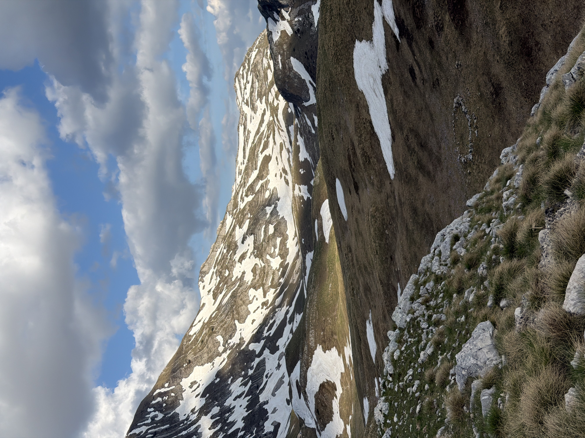 The image presents a mountainous landscape, possibly in the region of Montenegro, characterized by rocky terrain, patches of snow on the slopes, and a partially cloudy sky. The mountain peaks are sharply defined, with areas of both light and shadow highlighting the rugged texture of the rock formations. Vegetation is sparse, consisting mainly of grasses and small shrubs clinging to the rocky terrain. There are no people visible in the scene.