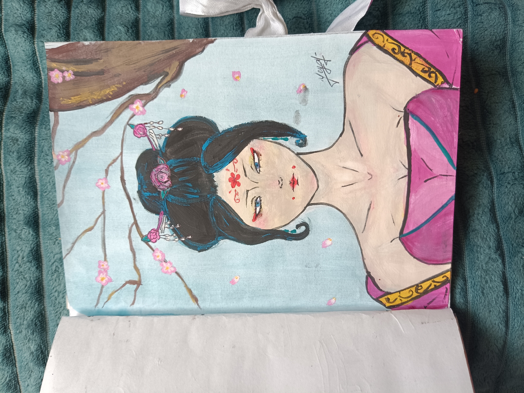 The image shows a painting of a woman, likely a geisha or similar figure, rendered in an amateurish style. She has black hair styled with pink floral ornaments and light blue accents. Her face is pale with red makeup details, including a flower on her forehead. She wears a pink garment trimmed with yellow. To her left is a tree branch with pink blossoms. The background is a light blue. The painting appears to be on a tote bag or a similar canvas-like material, which is partially opened, revealing white paper inside. The background of the photograph shows a textured, turquoise fabric. There is no location in the city of Posad, Russia.