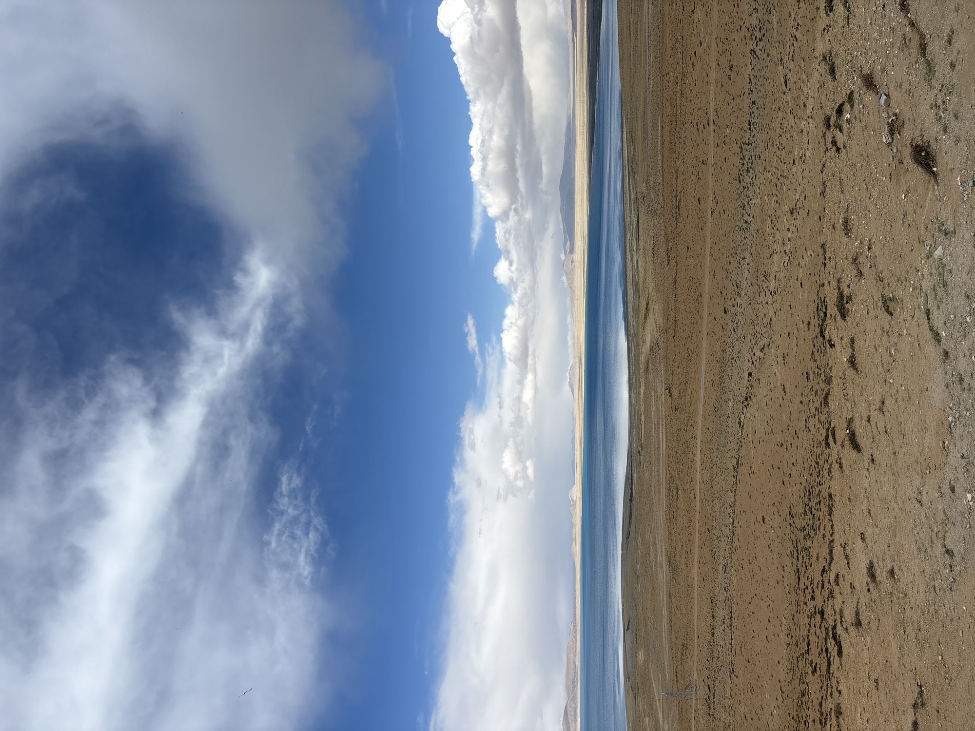 A landscape shot oriented vertically features a broad expanse of sky on the left, transitioning to a body of water and then dry, barren land on the right. The sky is predominantly blue with white clouds. A large, dense cloud formation is visible. The water stretches long and narrow, appearing bluish-grey. The land consists of rough, brown, arid terrain. The location is Gyirong County, China.