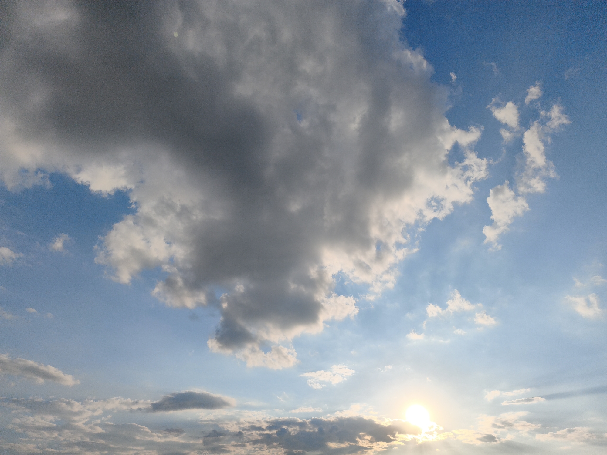 This is a photograph of the sky over Yemelivka, Ukraine, at sunset. The image is dominated by a large, dark gray cloud mass in the upper left quadrant, with smaller, wispy white clouds scattered across the bright blue sky. The sun is setting on the right side of the image, appearing as a bright, glowing orb with visible rays of light emanating outwards, illuminating the lower portion of the sky with a warm, golden hue. The lower horizon is obscured by clouds and the low angle of the sun. There are no people or man-made structures visible in the frame.