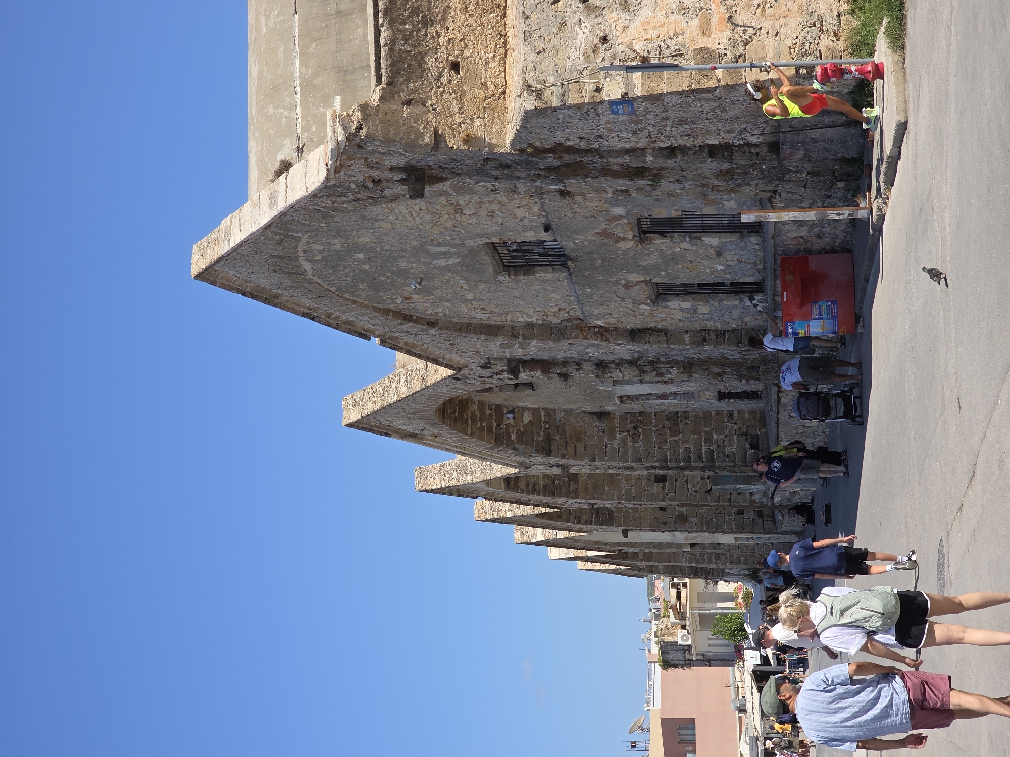 The image depicts a street scene in Chania, Greece, likely near the Venetian Harbor. In the foreground, a couple walks away from the camera, with the man wearing a striped blue and white shirt and maroon shorts, and the woman in a white top, black shorts, and a light green backpack. Behind them, other pedestrians are visible, including a child in a blue cap and shorts, and a man in a dark blue shirt.
The dominant feature of the background is a long, weathered stone building with a series of arched openings, characteristic of Venetian architecture found in Chania. The building has a rough, textured surface and appears to be quite old, with some signs of decay. The roofline is composed of a series of triangular gables.
To the right of the stone building, a man in a bright yellow vest and red shorts is performing a leg exercise, leaning against a red fire hydrant and a lamppost. A red metal structure, possibly a cooler or display, is situated near the building entrance.
Further in the background, other buildings, including a pale pink structure with satellite dishes on the roof, and smaller residential buildings, are visible. The sky is clear and bright blue. The street itself is paved and appears to be a public walkway or thoroughfare. A pigeon is visible on the street in the lower right portion of the image. The overall impression is a sunny day in a historic coastal city.