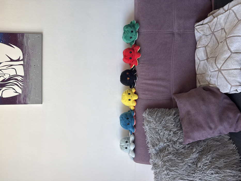 The scene depicts an interior setting in Plovdiv, Bulgaria, featuring a section of a purple upholstered sofa against a plain white wall. Six reversible octopus plush toys are arranged in a linear formation on the backrest of the sofa, from left to right: light blue, teal, yellow, black, red, and green. Each plush toy displays its "happy" or "neutral" facial expression. Resting on the sofa cushions are three distinct items: a large, fluffy grey pillow on the left, a smaller, smooth dark purple pillow in the center, and a light-colored blanket or throw with a repeating hexagonal pattern folded on the right. Above the sofa, a framed piece of artwork is mounted on the white wall. The artwork features a stylized, abstract illustration of a figure, predominantly in white, set against a background of purple and grey tones. No people are visible in the image.