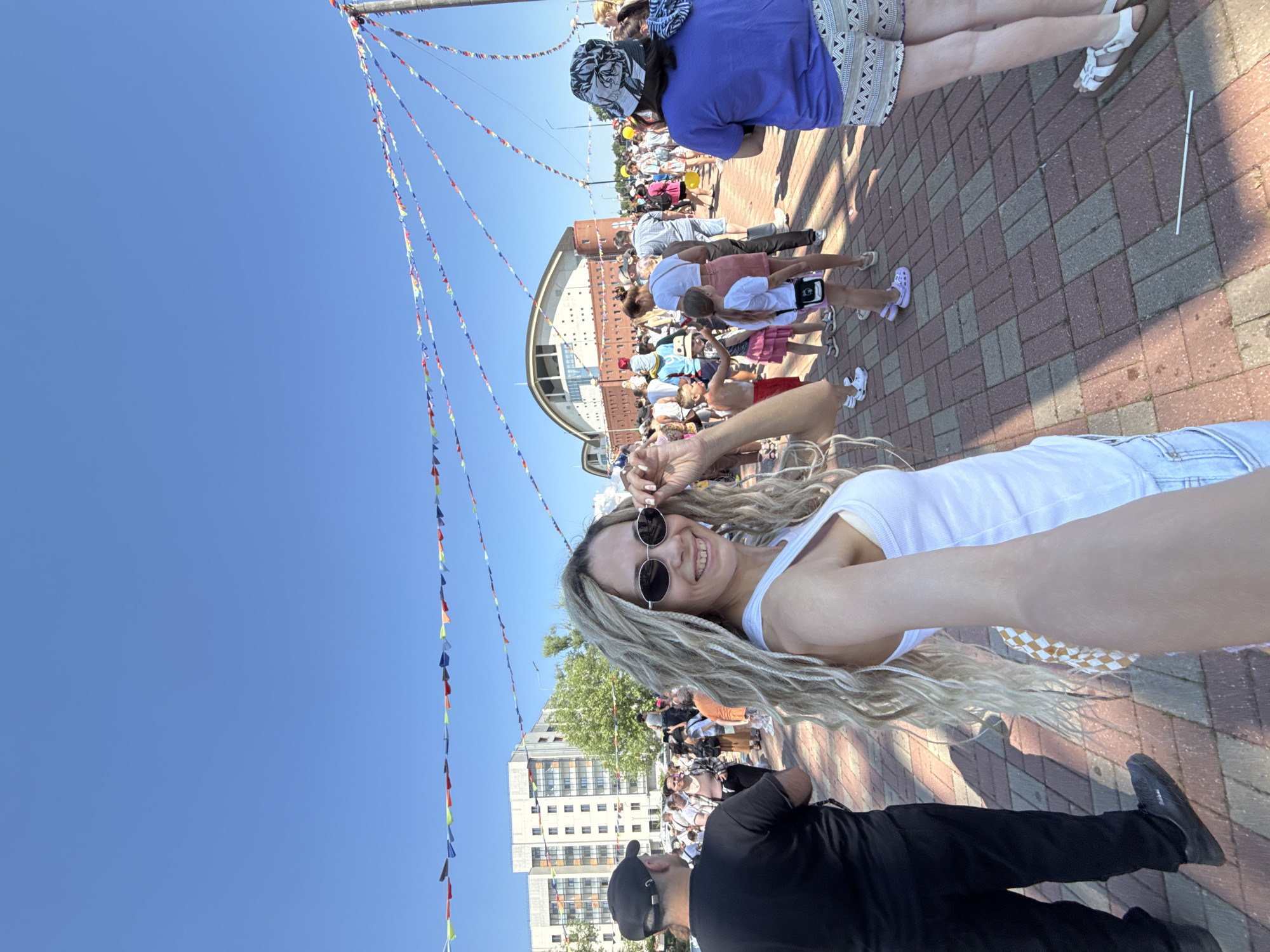 A woman with long, blonde braided hair and sunglasses is taking a selfie in an outdoor public space. She is wearing a white tank top and denim shorts. She is smiling and holding her sunglasses with her right hand. In the background, there are many people, a large brick building with a curved roof, and residential buildings. Colorful triangular flags are strung across the sky, and the ground is paved with brick. The scene appears to be a festival or event on a sunny day in the city of Zavolzhye, Russia.