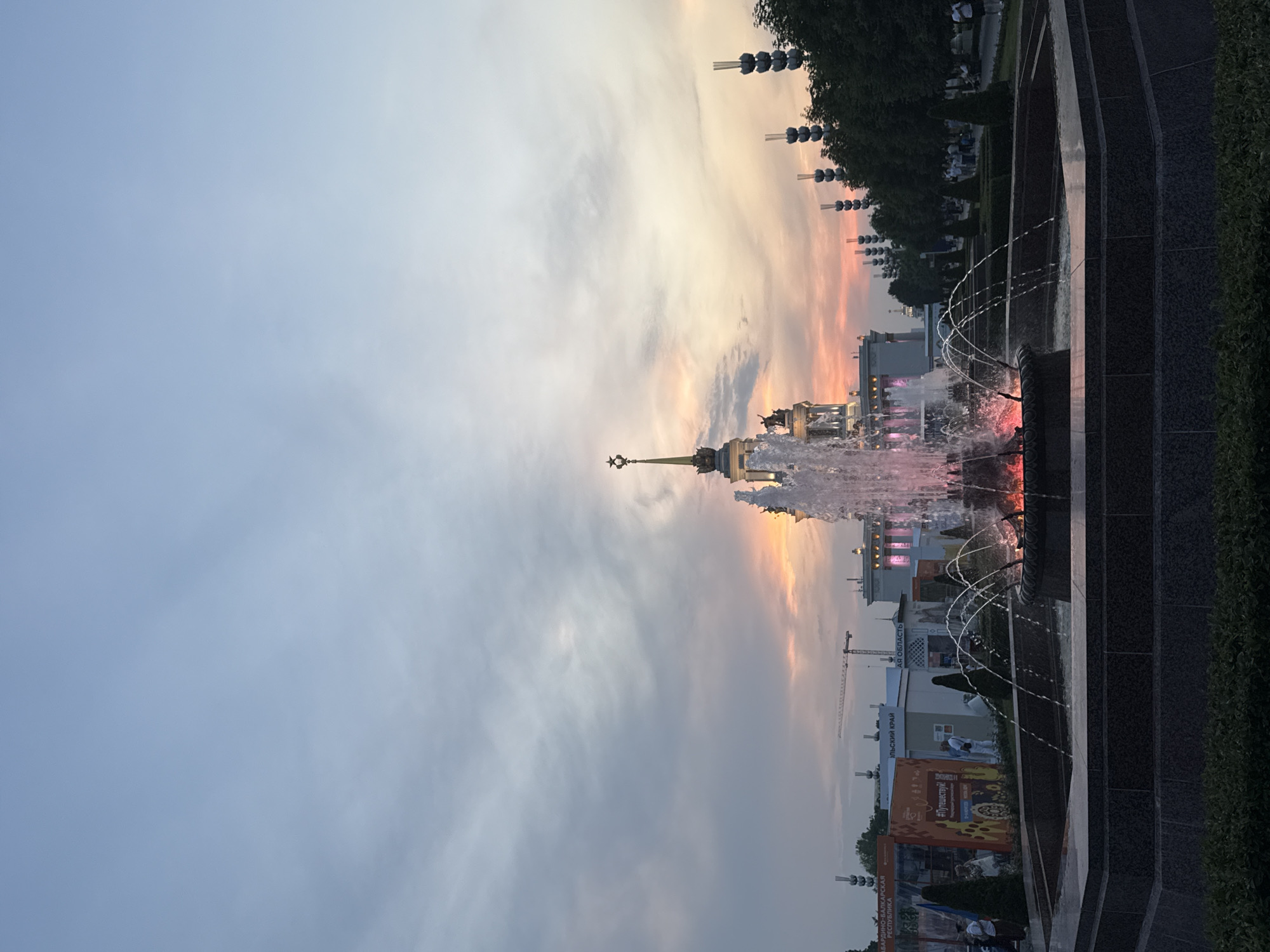 The image depicts a fountain in the foreground with water spraying upwards and outwards, illuminated by pink and red lights from below. Behind the fountain, a grand, ornate building with a star-topped spire stands prominently, bathed in the soft, warm hues of a sunset. The sky above is a blend of soft blues and greys, with hints of orange and pink near the horizon, indicating the time of day is late afternoon or early evening.
To the left of the main building, there are several exhibition booths or pavilions, one of which has signage with Russian text and images, suggesting a fair or exhibition. Some people are visible walking around these booths and in the background. To the right of the main building, a row of trees lines a pathway where more people are strolling. The overall scene suggests a public space, likely a park or exhibition ground, in Moscow, Russia, given the architectural style and the presence of Cyrillic script. The location is likely VDNKh (Exhibition of Achievements of National Economy) in Moscow, a popular park and exhibition center known for its impressive pavilions and fountains.
