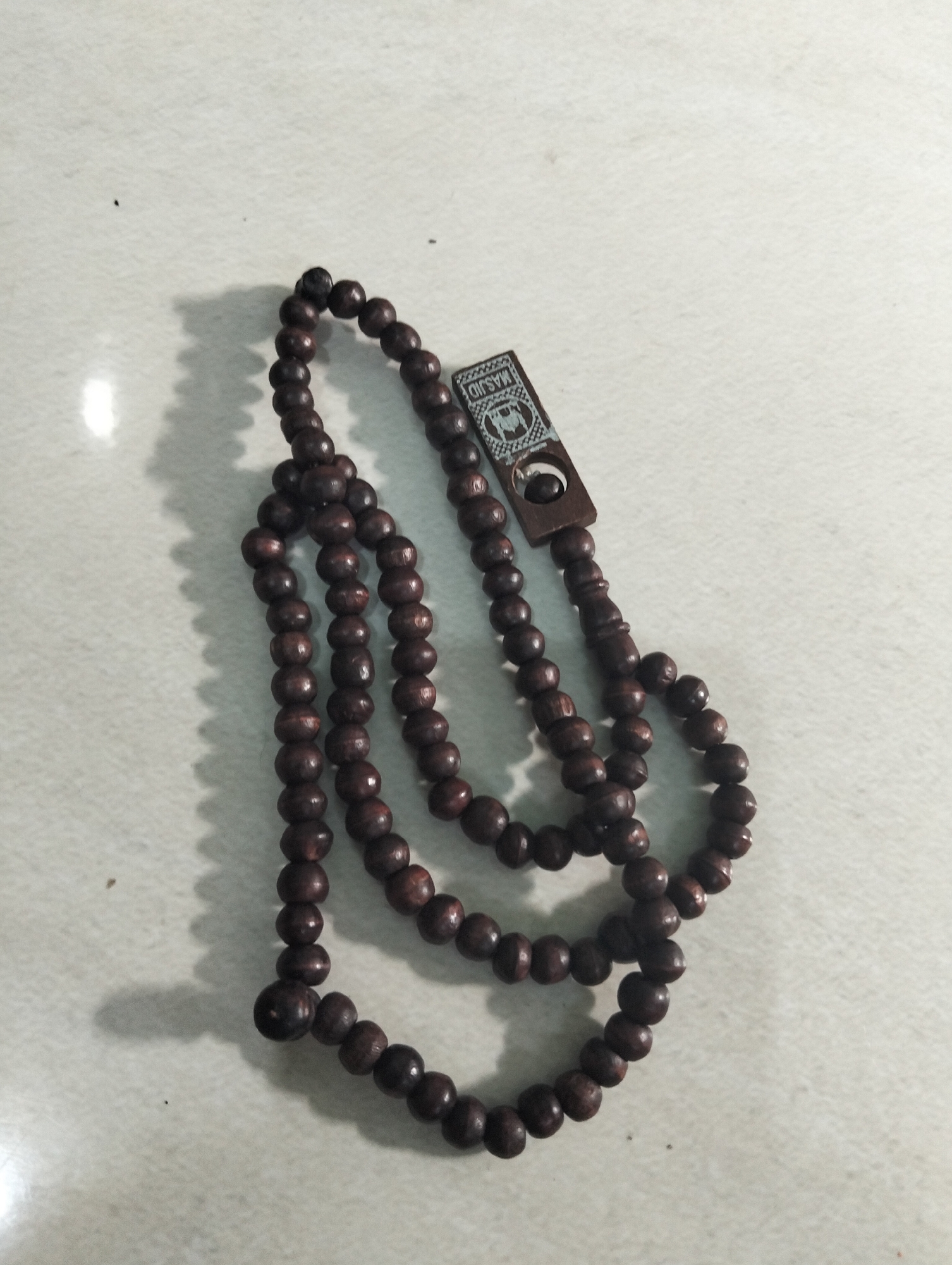 A close-up, overhead shot shows a set of dark brown prayer beads resting on a speckled white surface. The beads are uniform in size and shape, strung together in multiple loops. A small, rectangular wooden charm with carved Arabic script and an emblem is attached to the top strand of beads. The lighting casts soft shadows to the left of the beads, suggesting a light source from the right. The image does not depict any specific location within the city of Paciran, Indonesia.