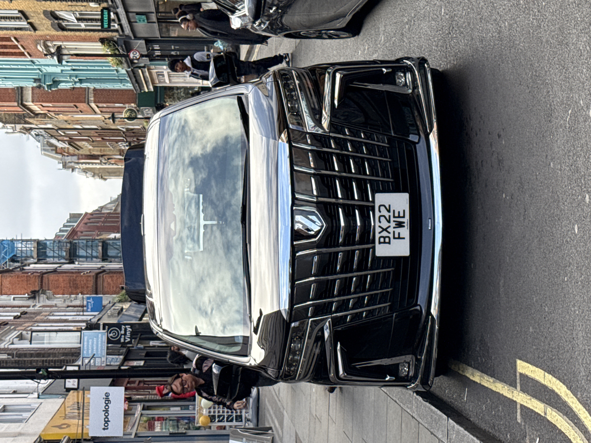 The image shows the front of a black car with a prominent chrome grille and the license plate "BX22 FWE." The car is parked on a street with double yellow lines. Reflections of clouds are visible in the windshield. Along the side of the street, a row of buildings is visible, including businesses with signage like "topologie" and "We Buy Vinyl." Several pedestrians are visible in the background. The buildings are typical of London architecture, suggesting the image was taken in the city of London, United Kingdom.