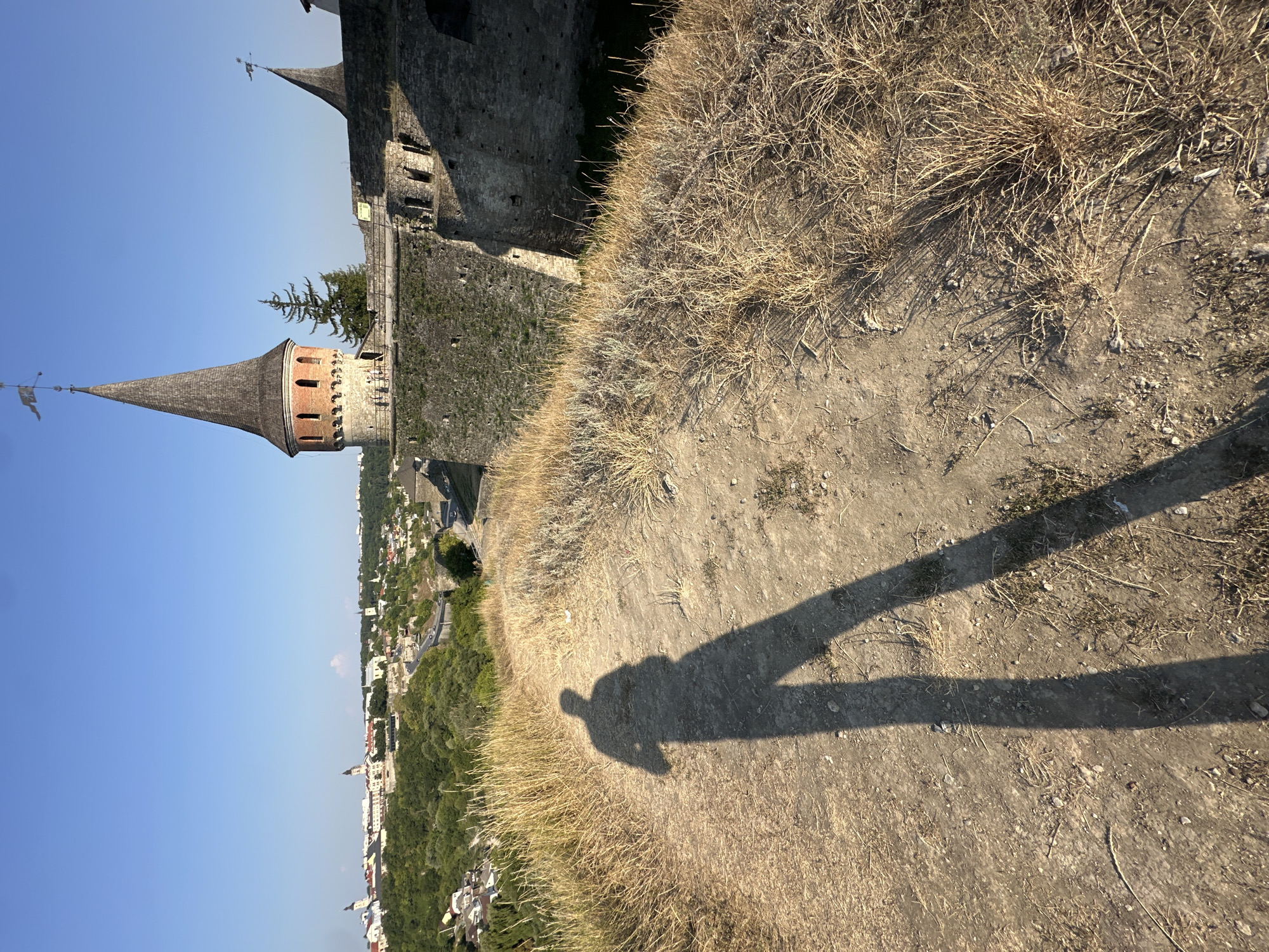 The image captures a view from a hill in Kamianets-Podilskyi, Ukraine, looking towards the historic Kamianets-Podilskyi Castle. In the foreground, a long shadow of a person is cast on a dry, dusty path with sparse, yellowed grass. The path curves towards the right. Behind the path, a weathered stone wall of the fortress is visible, constructed from large, irregular stones. To the left and in the background, a panoramic view of the city unfolds under a clear blue sky. Several buildings with red-tiled roofs and church spires are scattered across the landscape, nestled among green trees and undulating terrain. A prominent conical tower with a weathered wooden roof, part of the castle complex, stands tall against the sky, adorned with a flag at its peak. Another smaller, similar turret is visible to its right. The overall scene suggests a bright, sunny day, likely in the late afternoon due to the long shadows.