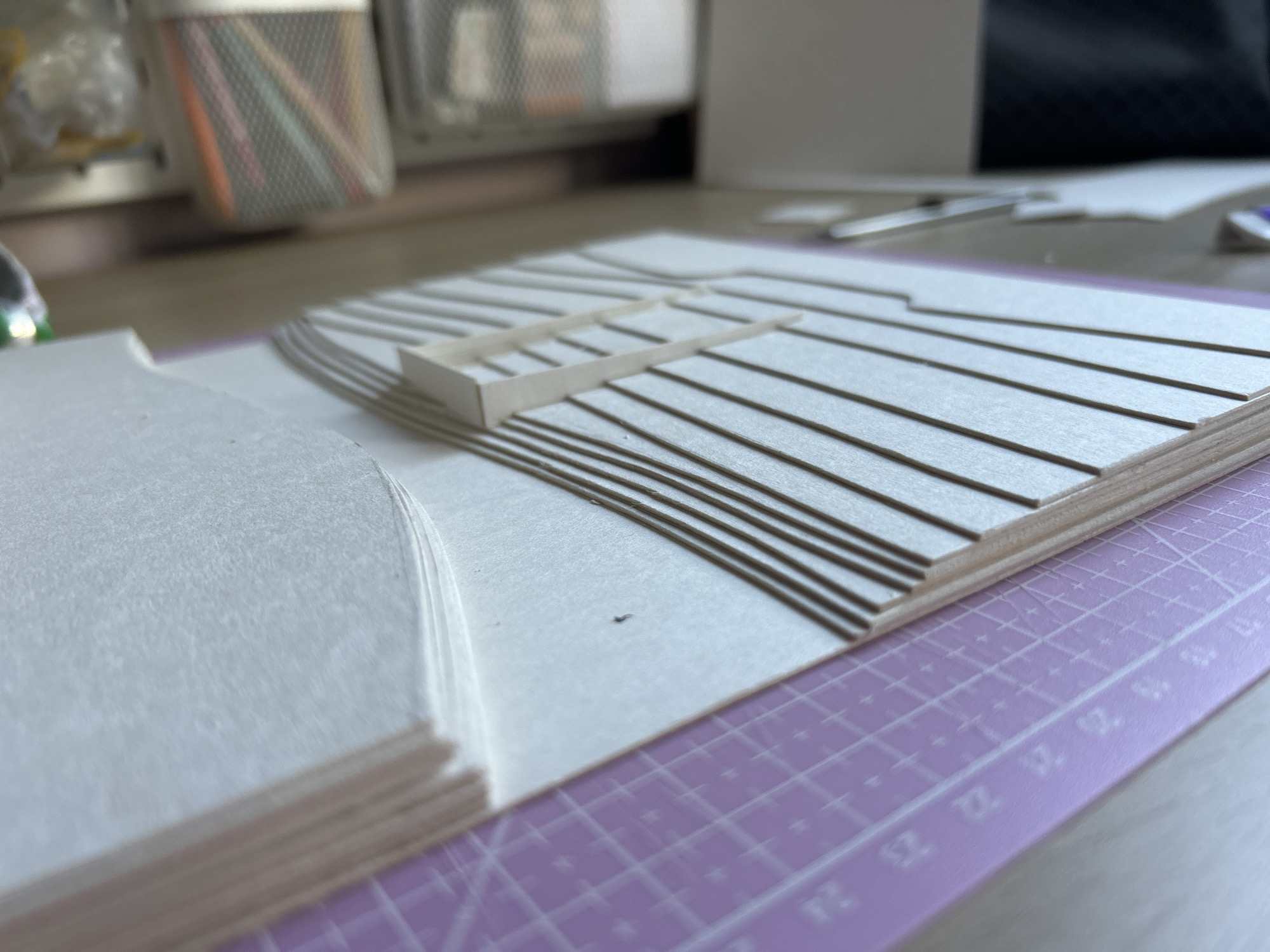 The image shows an architectural model of a multi-level structure, likely a house or a building, being constructed on a desk. The model is made of light-colored paper and thin wooden strips. The base of the model features several layered horizontal platforms with varying heights, creating a stepped effect. A rectangular structure, possibly representing a room or a section of the building, is placed on one of these platforms. The model is situated on a purple cutting mat with grid lines and measurements, indicating a crafting or prototyping activity. In the background, out of focus, are art supplies like pencils in a container and some paperwork. The lighting suggests an indoor environment, possibly a workshop or a studio. The scene is devoid of any people or immediate interaction. The location in Sofia, Bulgaria, is not discernible from the visual information provided in the image.