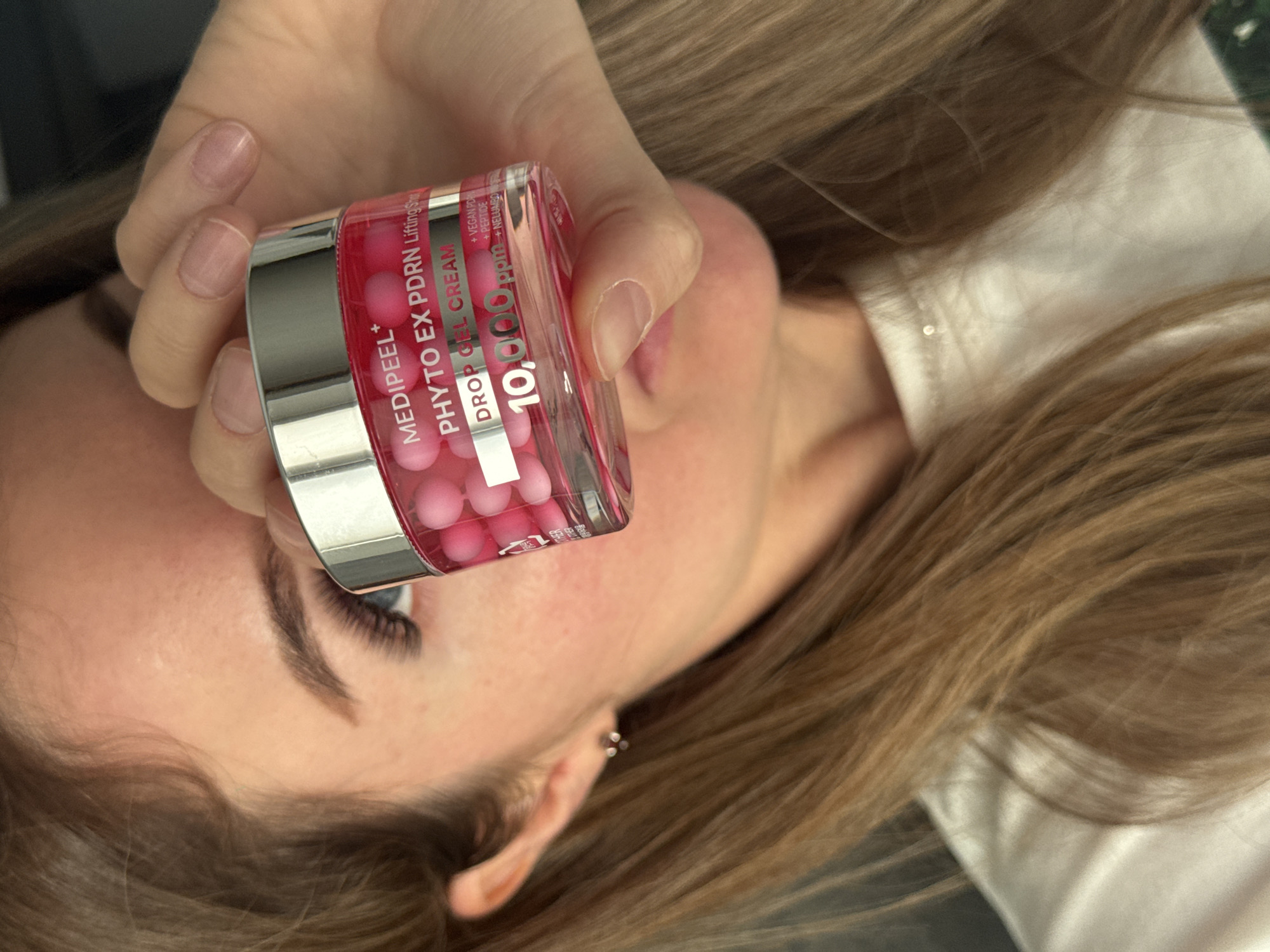 A person holds a jar of Medipeel Phyto Ex PDRN Lifting Shot Drop Gel Cream in front of their face. The jar contains pink spheres and is labeled with the product name and "10,000 ppm". The person's eye, eyebrow, and part of their hair are visible. The background is blurred and appears to be indoors.