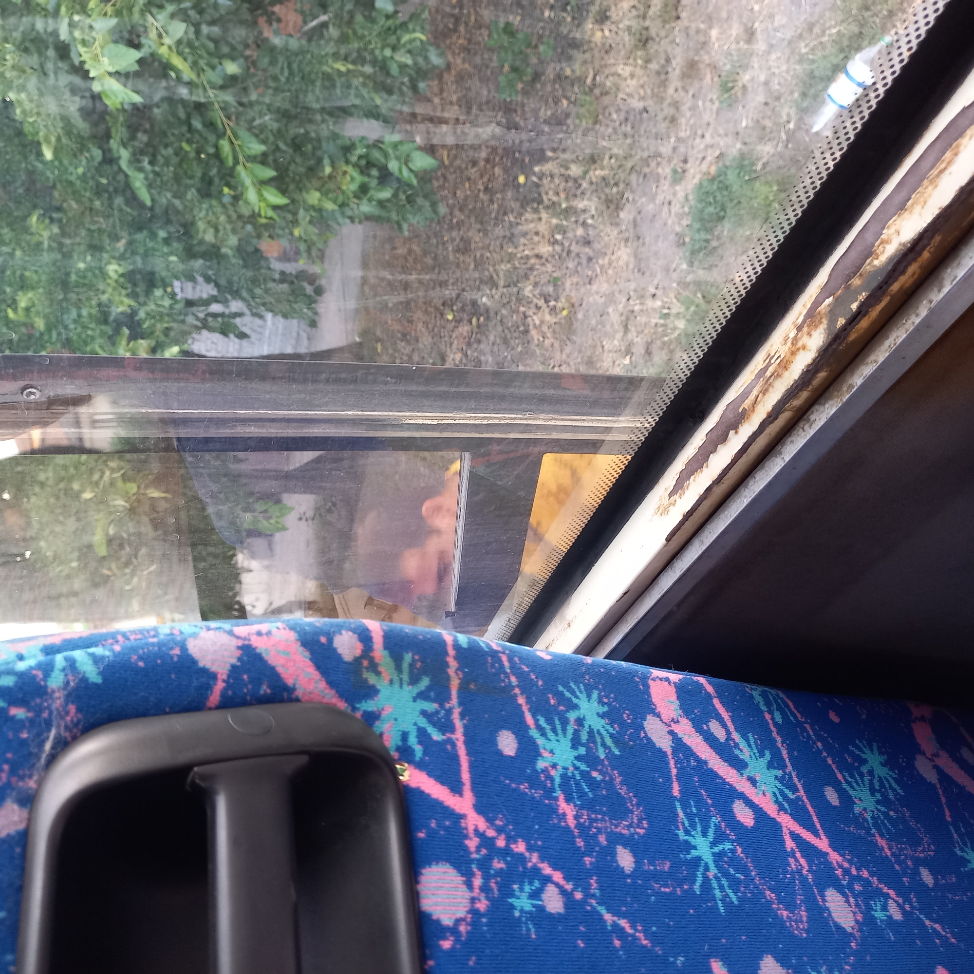The image shows the interior of a bus, with a blue seat patterned with pink and light blue splatters in the foreground. To the right is a window looking out onto a moderately vegetated area with trees and dry grass. A person's reflection is visible in the window. The window frame shows signs of rust. The location is specified as Kropyvnytskyi, Ukraine.