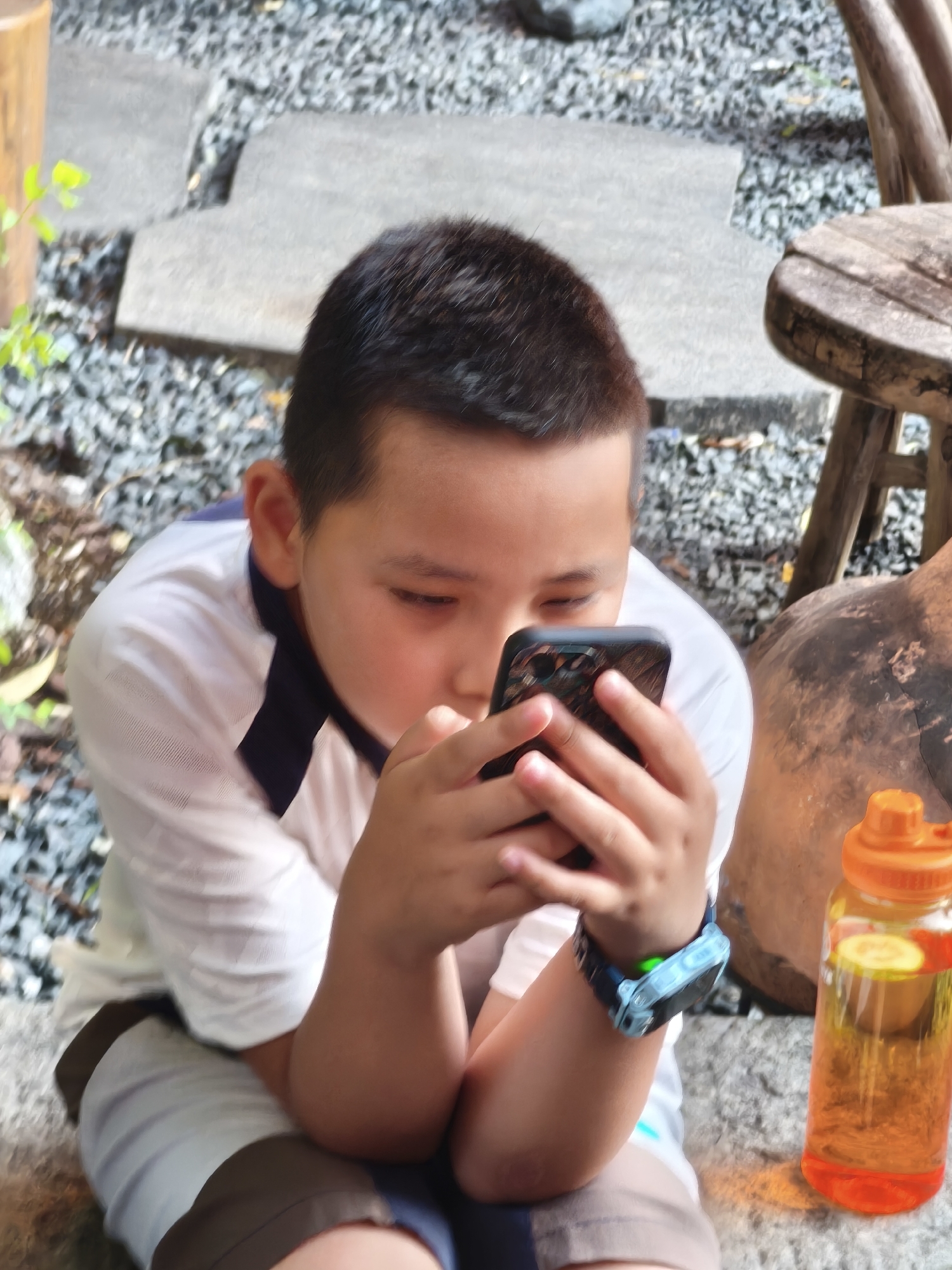 A young boy, with short dark hair, is focused on a smartphone held in his hands. He is wearing a white polo shirt with navy trim and shorts. On his left wrist, he wears a blue digital watch with a glowing green light. Next to him, on the right, is a clear plastic water bottle with an orange cap, containing a yellow liquid with slices of lemon or lime and possibly other fruit. The boy appears to be sitting outdoors on a surface covered in small gray stones, with larger flat gray stones and some greenery visible in the background. A rustic wooden stool is also present to the right of the boy, with a large, dark, rounded object behind the water bottle, which may be a decorative planter or sculpture. The overall scene suggests a relaxed outdoor setting, possibly a garden or patio area. The location is identified as Heping District, China.