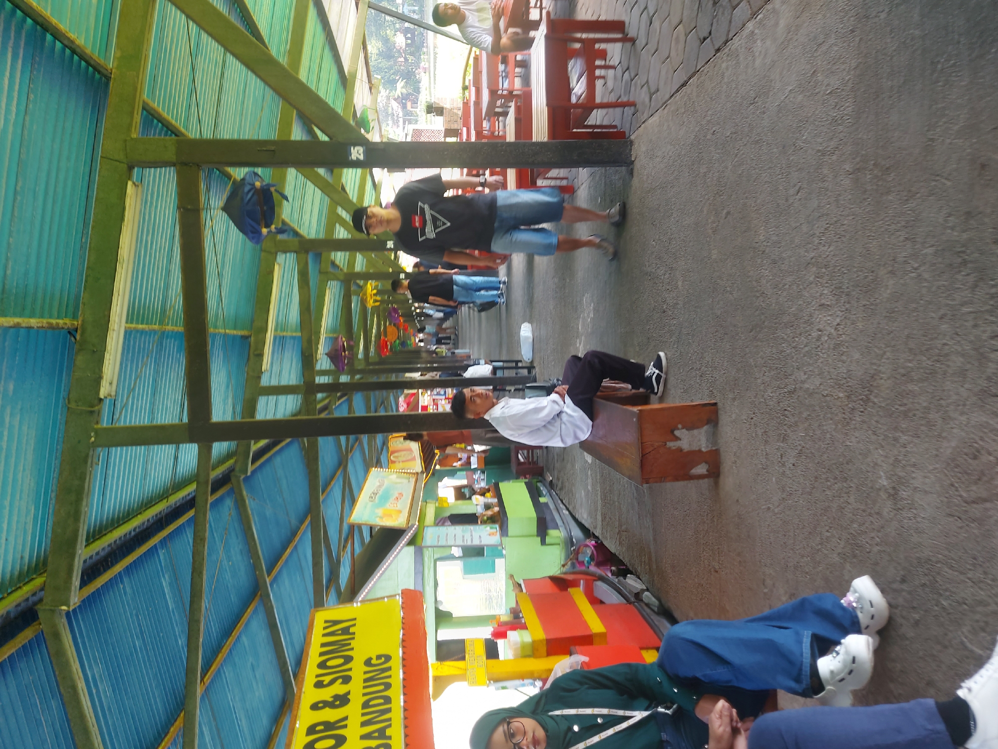 The image depicts a covered walkway at a food stall area, likely in Lembang, Indonesia, given the sign that reads "GOR & SIOMAY BANDUNG." The structure has a blue corrugated metal roof supported by green-painted metal beams. Various food stalls are visible, one on the left with a yellow sign displaying "GOR & SIOMAY BANDUNG" and red and yellow accents. Another stall to its right is green and appears to be selling drinks or other food items.
In the foreground, on the left, a person wearing a green headscarf and glasses is seated, with another person's legs visible to their left. Further down the walkway, a man in a white shirt and dark pants sits on a wooden bench. Across the walkway, a man in a black t-shirt, blue shorts, and a baseball cap walks towards the camera. Other people are visible in the background, including one person in a white t-shirt sitting at a red table. Red tables and chairs are arranged in an outdoor seating area to the right. Decorative lanterns, some colored blue and yellow, hang from the ceiling. The ground is paved with concrete. The overall impression is a casual outdoor dining or rest area.