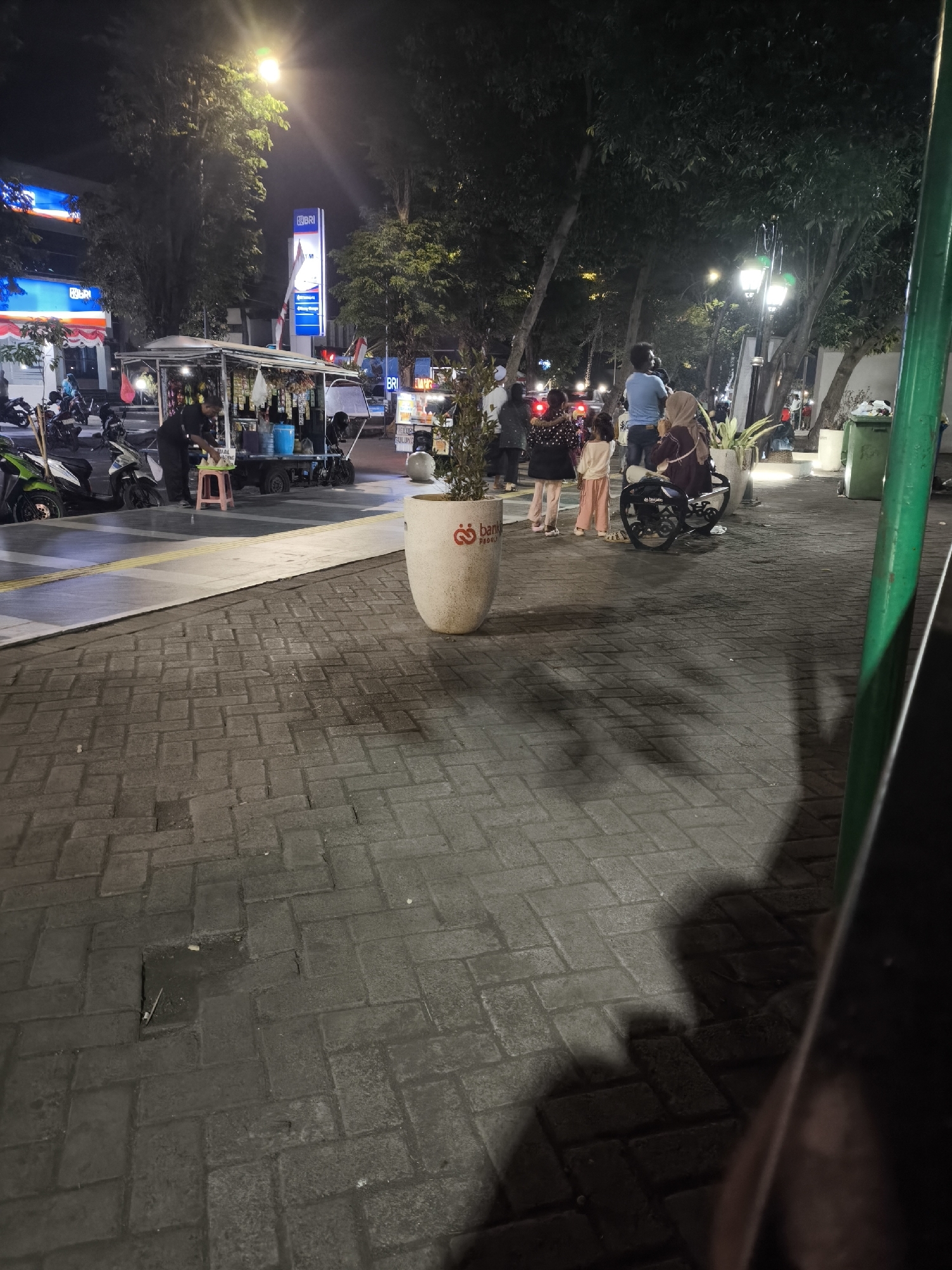 The image depicts a nighttime street scene in Lumajang, Indonesia. In the foreground, a paved walkway made of gray brick pavers is visible, with some sections appearing wet or discolored. A large, light-colored decorative pot with a logo is positioned on the walkway.
In the midground, a food stall on wheels is set up, featuring various items for sale and illuminated by lights. A man in a dark shirt and pants is attending to the stall, holding a tray. Several motorcycles are parked nearby. Further back, a group of people, including adults and children, are gathered. One person is sitting on a motorcycle.
The background reveals buildings with illuminated signs, including "BRI," a bank. Trees line the street, casting shadows on the walkway. Streetlights illuminate the scene, with one prominent lamppost with multiple lights casting a bright glow. The overall atmosphere suggests a lively evening in a public urban space.