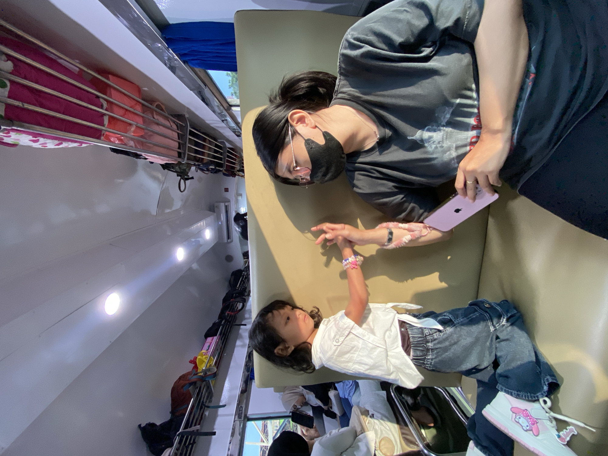 The image shows a woman wearing a black mask and glasses, lying on a beige cushioned seat. She is holding a pink phone and interacting with a young child, lying on the same seat. The child wears a white shirt and jeans, and has a bracelet on their wrist. The scene appears to be inside a train or similar passenger vehicle. Luggage is visible on overhead racks. The location is Kidul Dalem, Indonesia.