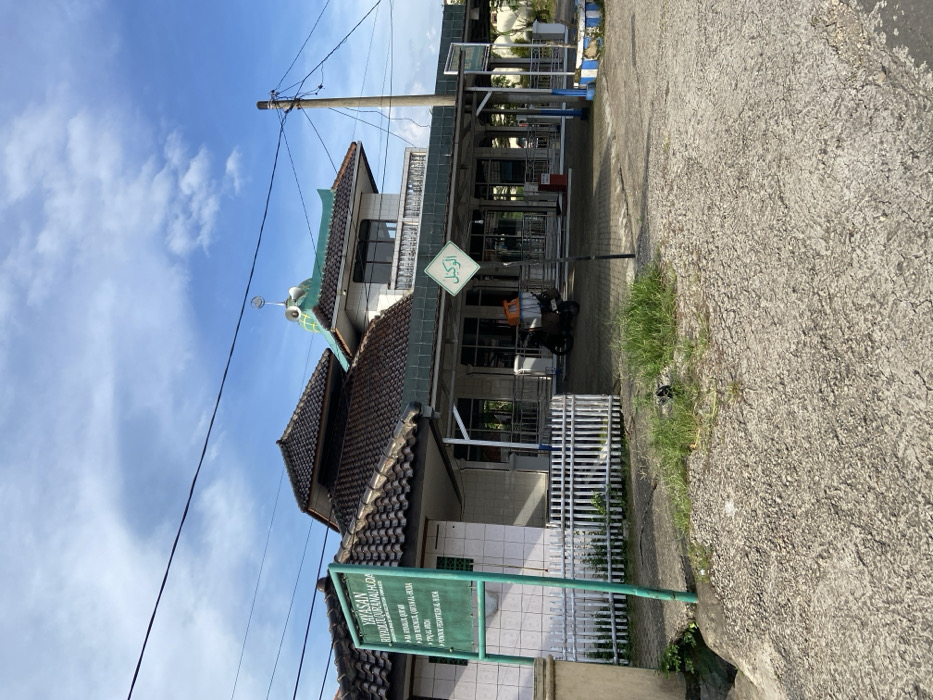 The image shows the exterior of a mosque in Sukatani, Indonesia, located at the coordinates -6.643 latitude and 107.415 longitude. The mosque has a white facade with a green tiled roof and a single minaret. There is a sign on the front of the mosque that reads "YAYSAN RIYADUL QUR'AN MA'AHIDA" in Indonesian, which translates to "Riyadul Quran Ma'ahida Foundation". The sign is located on a green pole in front of a white, wooden fence. The mosque has large windows and a balcony overlooking the street. The environment is urban and the street is paved with grey gravel. There are several power lines running overhead. A motorcycle is parked in front of the mosque. A sign with an arrow pointing to the right that says "الزكاة" in Arabic is located near the front of the mosque.