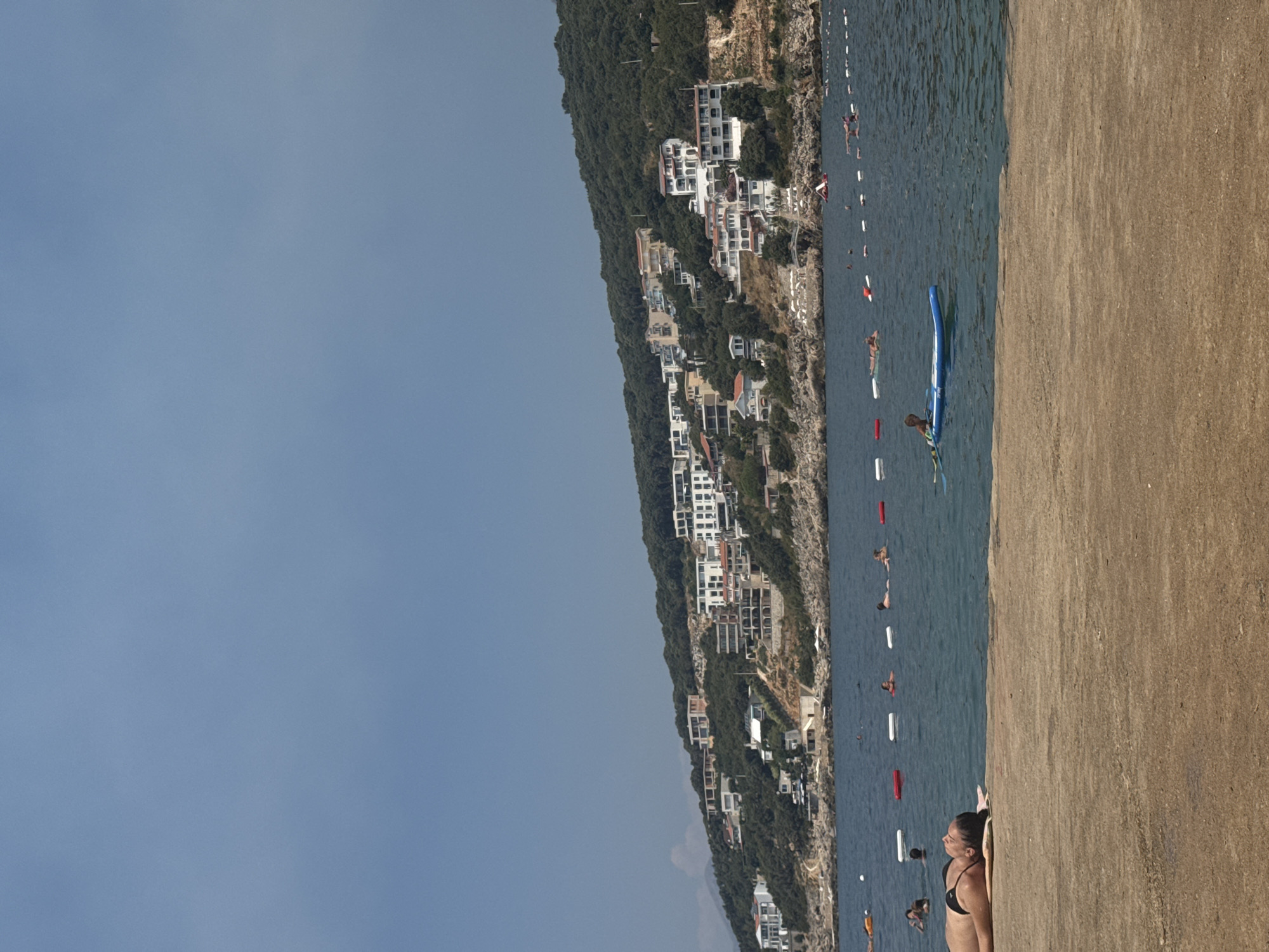 In the foreground, a person lies on their back on a concrete surface, facing left, with their eyes closed and hands resting on their chest. They are wearing a black bikini. The concrete surface extends to the left and right edges of the frame and occupies the bottom third of the image.
Beyond the concrete, a body of blue water stretches to the horizon. Several buoys, some white and some red, are visible in the water, marking a boundary. Numerous people are scattered throughout the water, some swimming, some floating, and some standing. One person is on a blue paddleboard, and another is on a lighter-colored inflatable raft.
In the background, a lush green hillside rises steeply from the water's edge. Dotted along the hillside are numerous white buildings with red or white roofs, characteristic of a coastal town. The architecture suggests a Mediterranean or Balkan style. The sky above is a clear, pale blue. The scene depicts a sunny day at a beach or coastal resort area. The location is identified as a beach in Kunje, Montenegro.