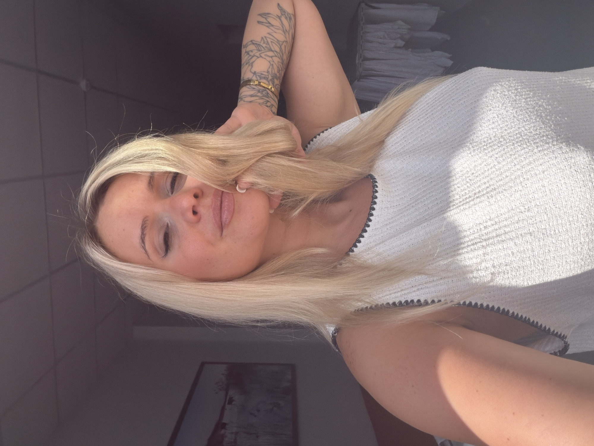The image shows a blonde woman taking a selfie. She is wearing a white sleeveless top with a black trim. Her right arm is raised, and her hand is in her hair. She has a tattoo on her left arm and a gold bracelet on her right wrist. The background is blurred, but it appears to be an indoor setting with a ceiling and a wall. A framed picture is visible on the wall to the left. The lighting suggests it is daytime. There is no identifiable information to place the image in Sarapul, Russia.