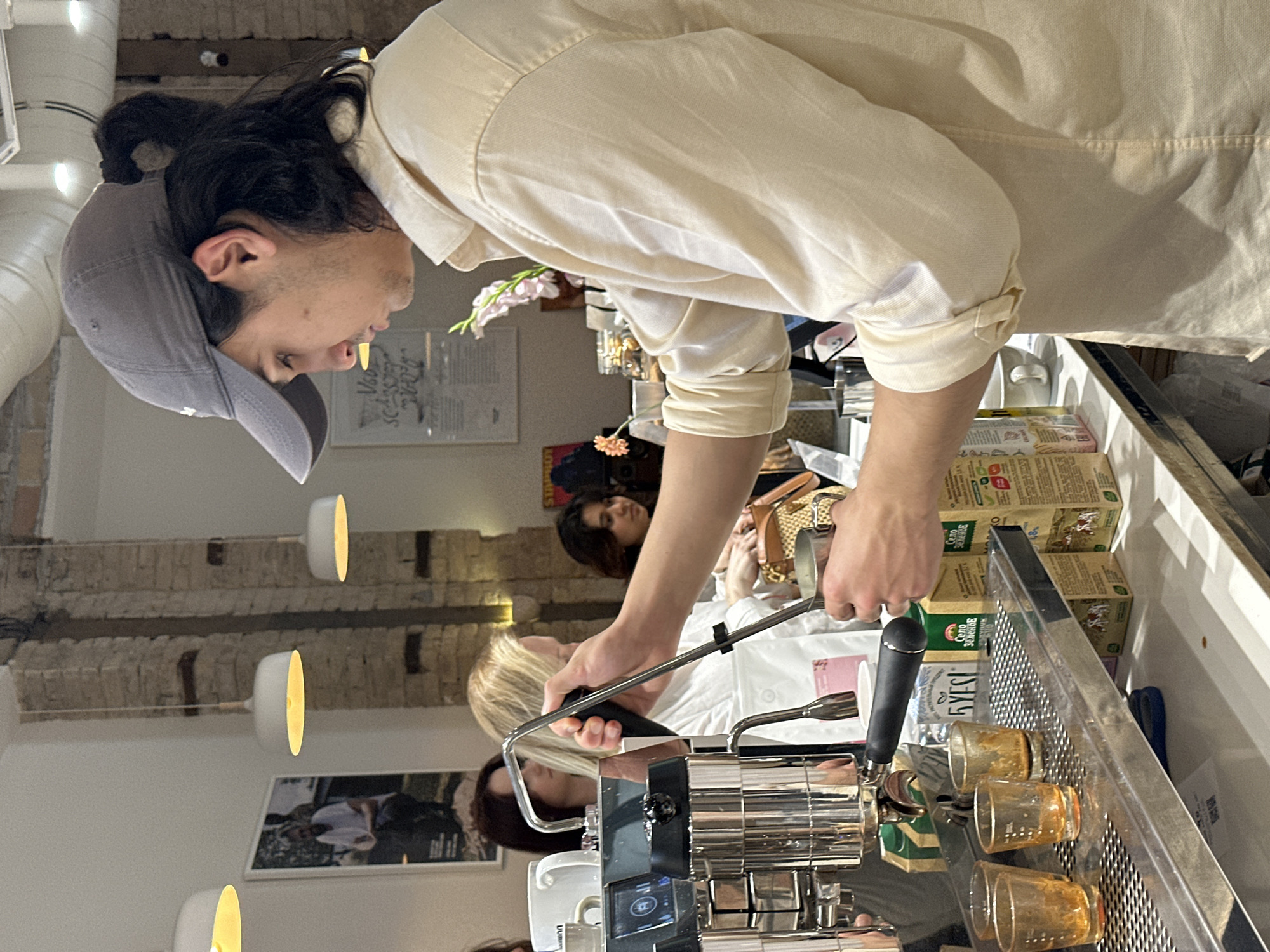 A barista in a tan button-down shirt and a gray baseball cap operates an espresso machine. Four small glass cups are on the machine's drip tray. Behind the barista, customers are seated at tables. The scene is indoors, with brick detailing on the wall and pendant lights overhead. Boxes of tea or coffee are stacked near the barista. The location is likely a cafe, possibly in Tashkent, Uzbekistan. The barista is pulling espresso shots for the customers.