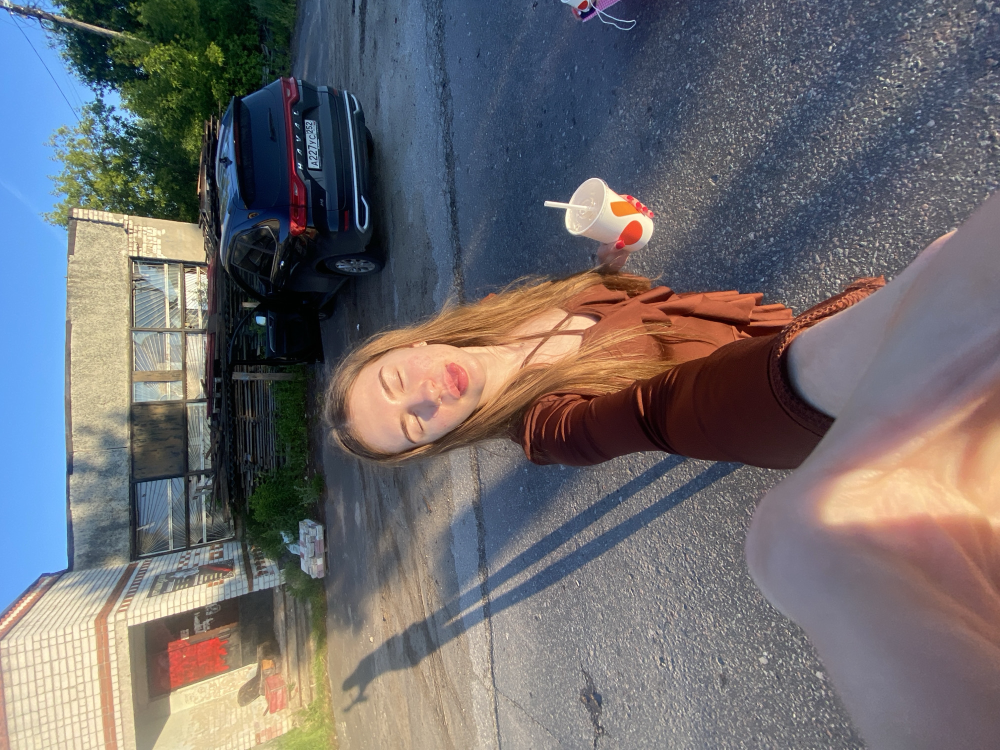 A young woman with long blonde hair, wearing a brown dress, is taking a selfie outdoors during the day in Medvevo, Russia. She is sticking her tongue out and holding a beverage cup with a straw in her right hand. Her left hand is also visible, close to the camera. A black Haval SUV with license plate A227YC 252 is parked behind her, with its driver's side door open. In the background, a light-colored brick building with a red door and dark windows is visible. There is a long shadow cast on the asphalt ground from the woman. Trees and foliage are present behind the building and the car. The sky is clear and blue.