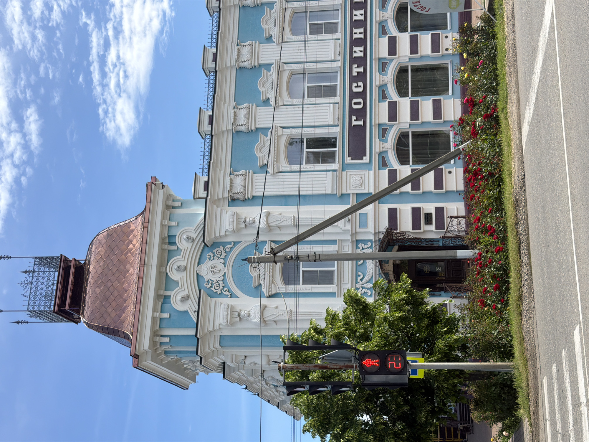 The image displays a vibrant street scene featuring a stately, multi-story building under a clear, bright blue sky with wispy white clouds. The building, likely a hotel given the text "ГОСТИНИЦА" (GOSTINITSA) written vertically on its facade, exhibits a light blue main color contrasted with elaborate white architectural details such as columns, cornices, and decorative window frames. A prominent feature is its unique copper-colored, diamond-patterned dome roof, topped with an intricate black wrought-iron railing.
At ground level, a lush green strip of land separates the building from the road, filled with dense bushes adorned with numerous red and some yellow roses. In the foreground, a pedestrian traffic light is visible, illuminated red with a standing figure icon and the digital countdown "24" seconds. Other vehicular traffic lights are mounted on the same pole. Black utility wires crisscross in front of the building. Beyond the flowery median, an asphalt road with white lane markings, including a solid white line and a dashed line, is present. Green trees and foliage are scattered throughout the scene, adding to the natural elements. The sunny conditions suggest it is daytime. The scene depicts a lively urban or town environment in Leningradskaya, Russia, blending architectural heritage with modern street infrastructure.