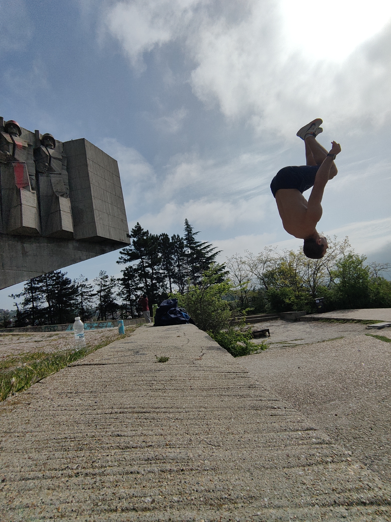 The image shows a man performing a backflip in front of a monument in Varna, Bulgaria. The monument is a large, concrete structure with abstract figures. The man is shirtless and wearing dark shorts and sneakers. He is in mid-air, with his head and torso upside down. The sky is partly cloudy. In the foreground, there is a concrete surface with grass and some plants growing along the edge. There is a water bottle and other objects on the concrete surface. In the background, there are trees and greenery.